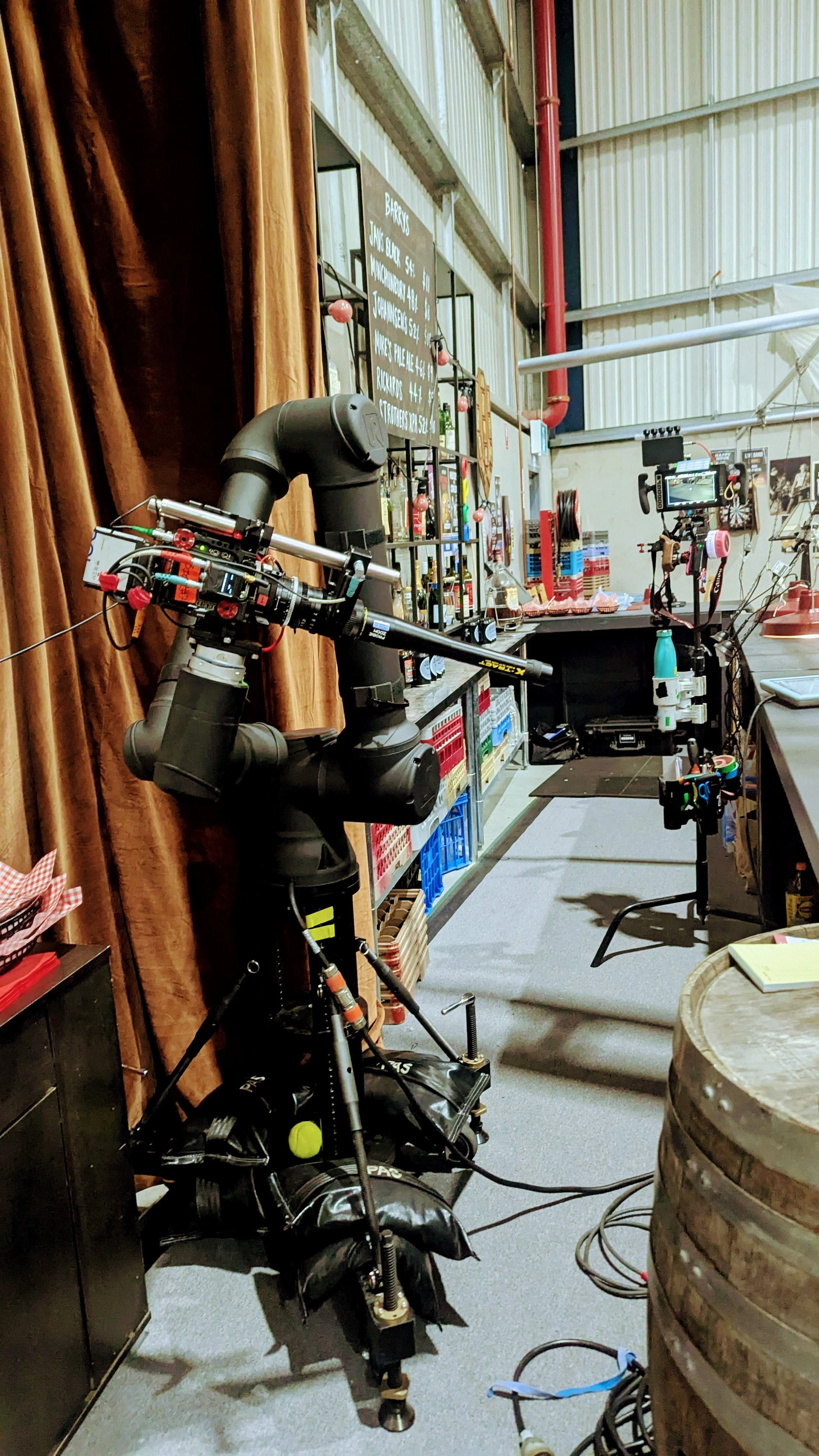 A robotic arm setup inside a bar or restaurant with a chalkboard menu in the background and various equipment and supplies around.