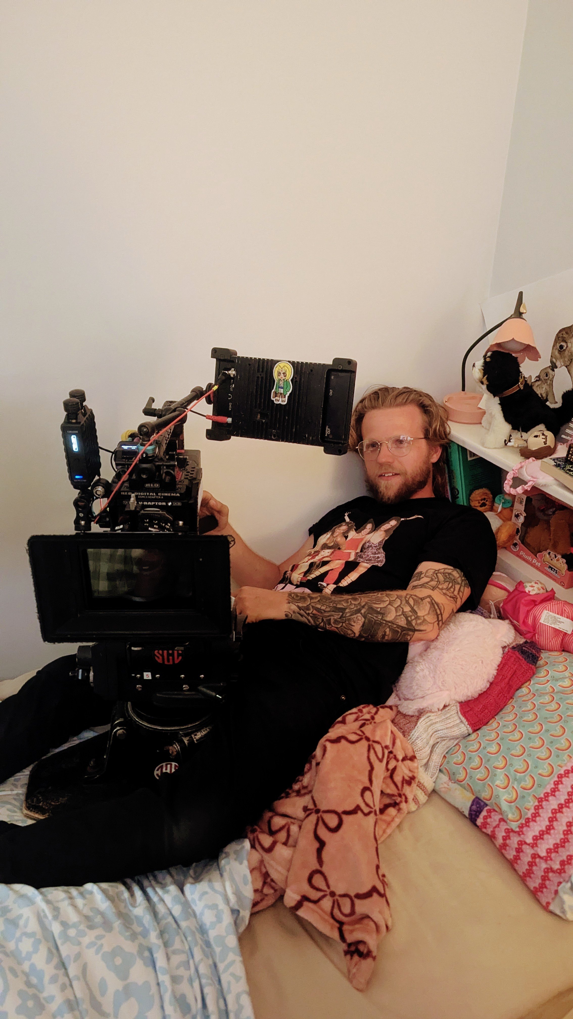 A man with glasses, tattoos, and wavy blonde hair is lying on a bed with a camera mounted on a tripod, filming or taking a picture. The bed is covered with colorful blankets, and there are various stuffed animals and toys on a nearby shelf.