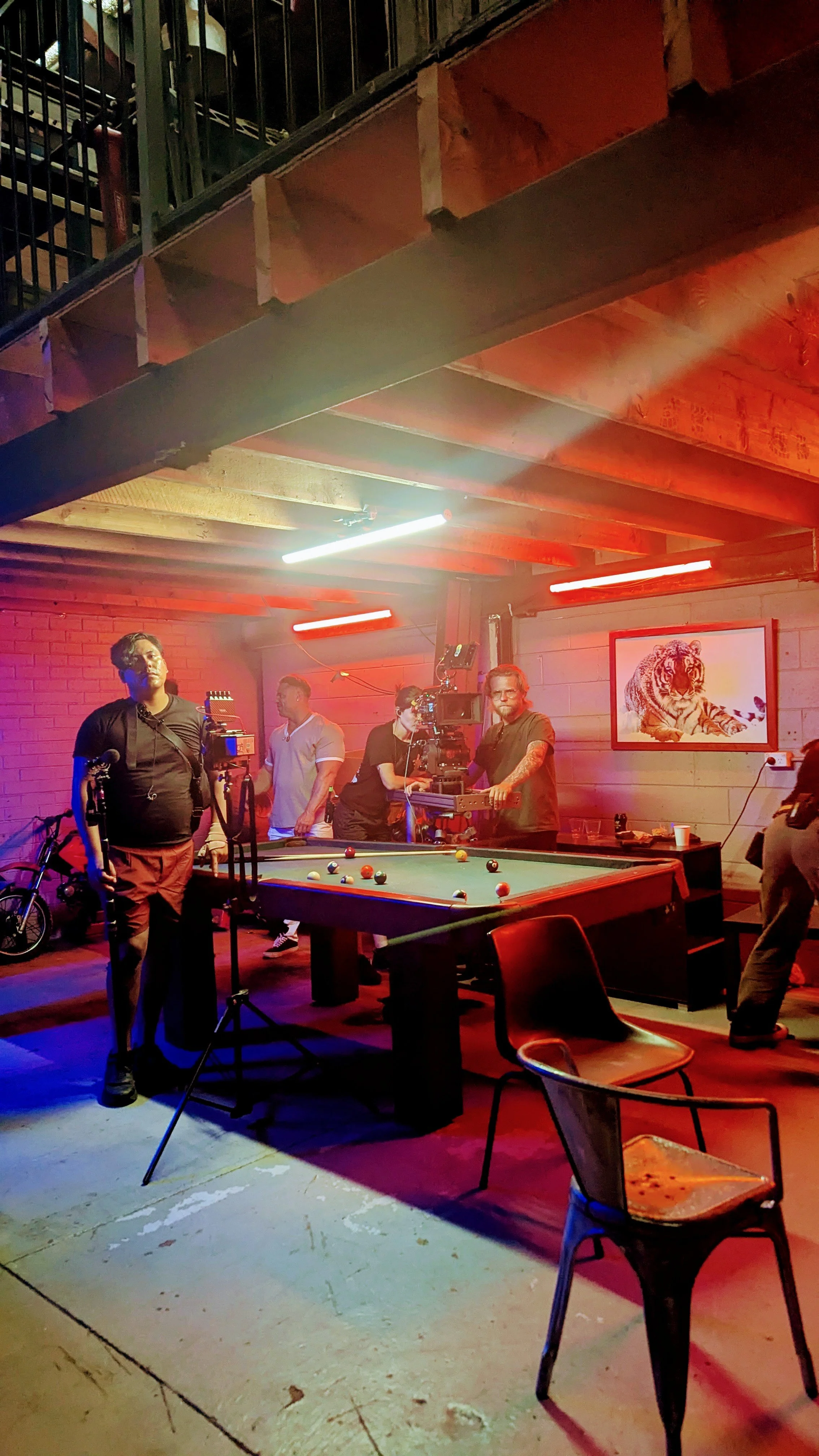 A group of people setting up filming equipment around a pool table in a room with red and blue lighting, brick wall, and a picture of a tiger on the wall.