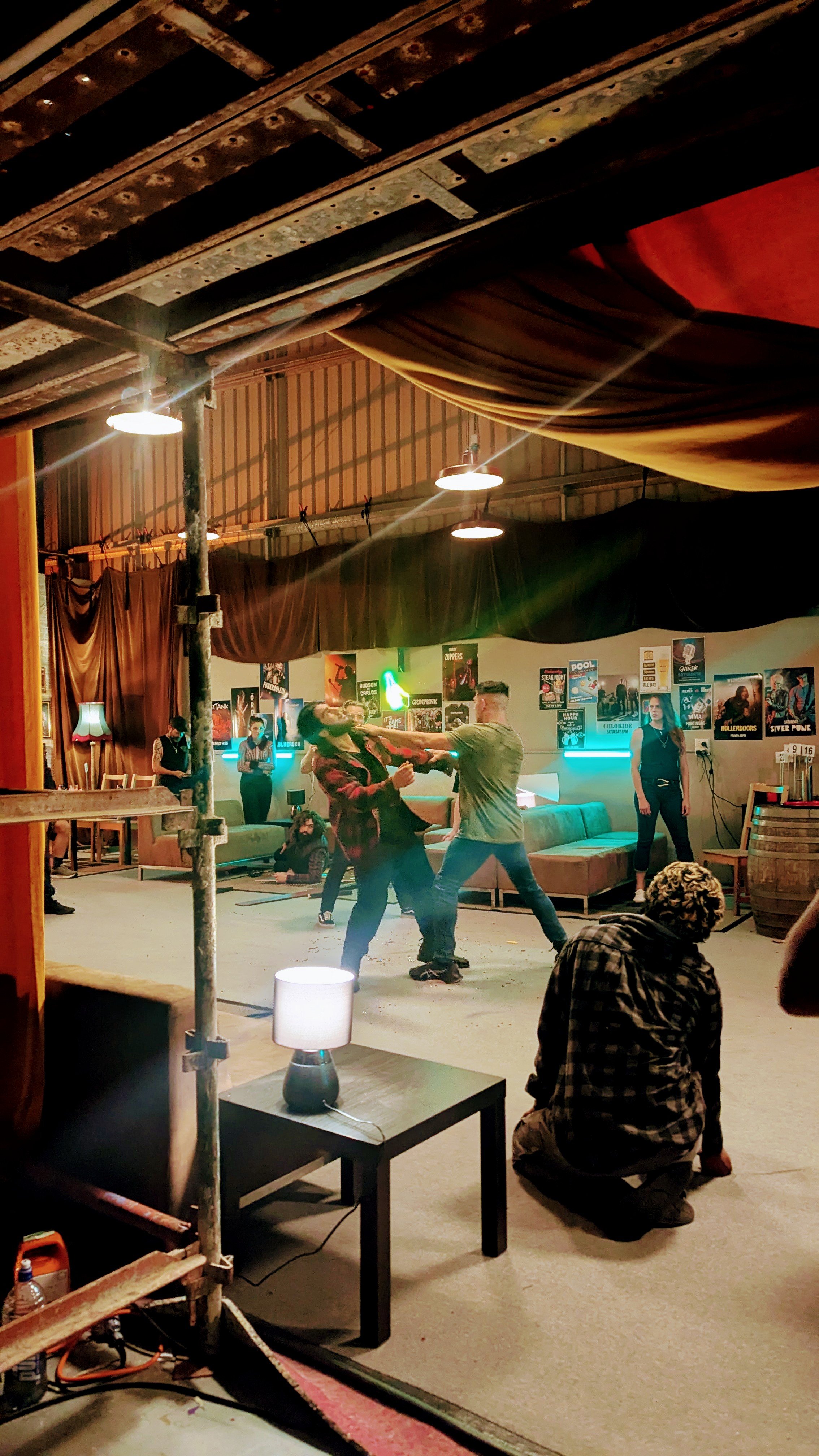 People dancing in a rustic indoor venue with posters on the wall, colorful lighting, and casual furniture, indicating a lively social gathering or party.