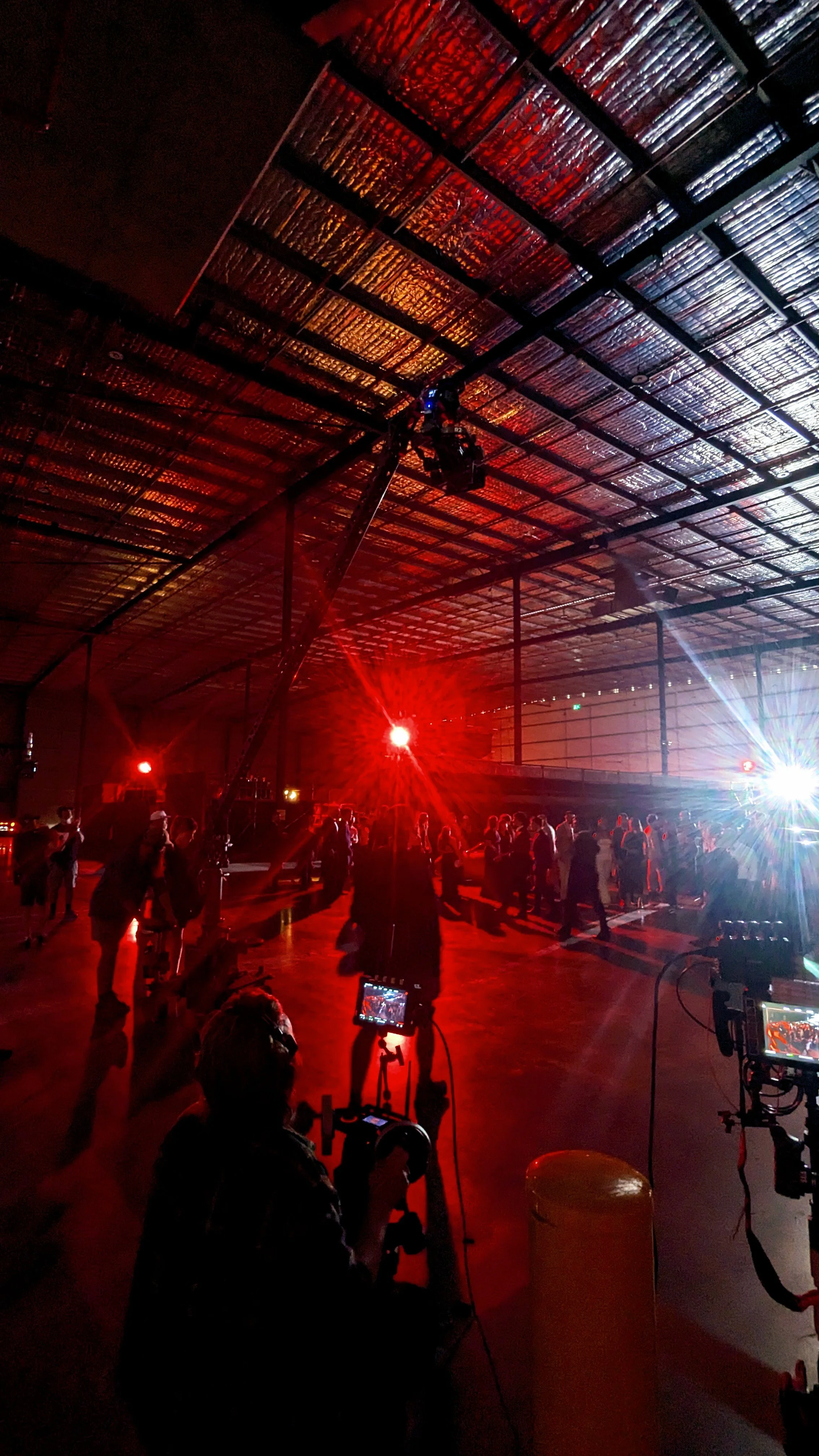 People dancing in a warehouse with red and white lights, seen from behind the camera operator with two cameras on tripods.