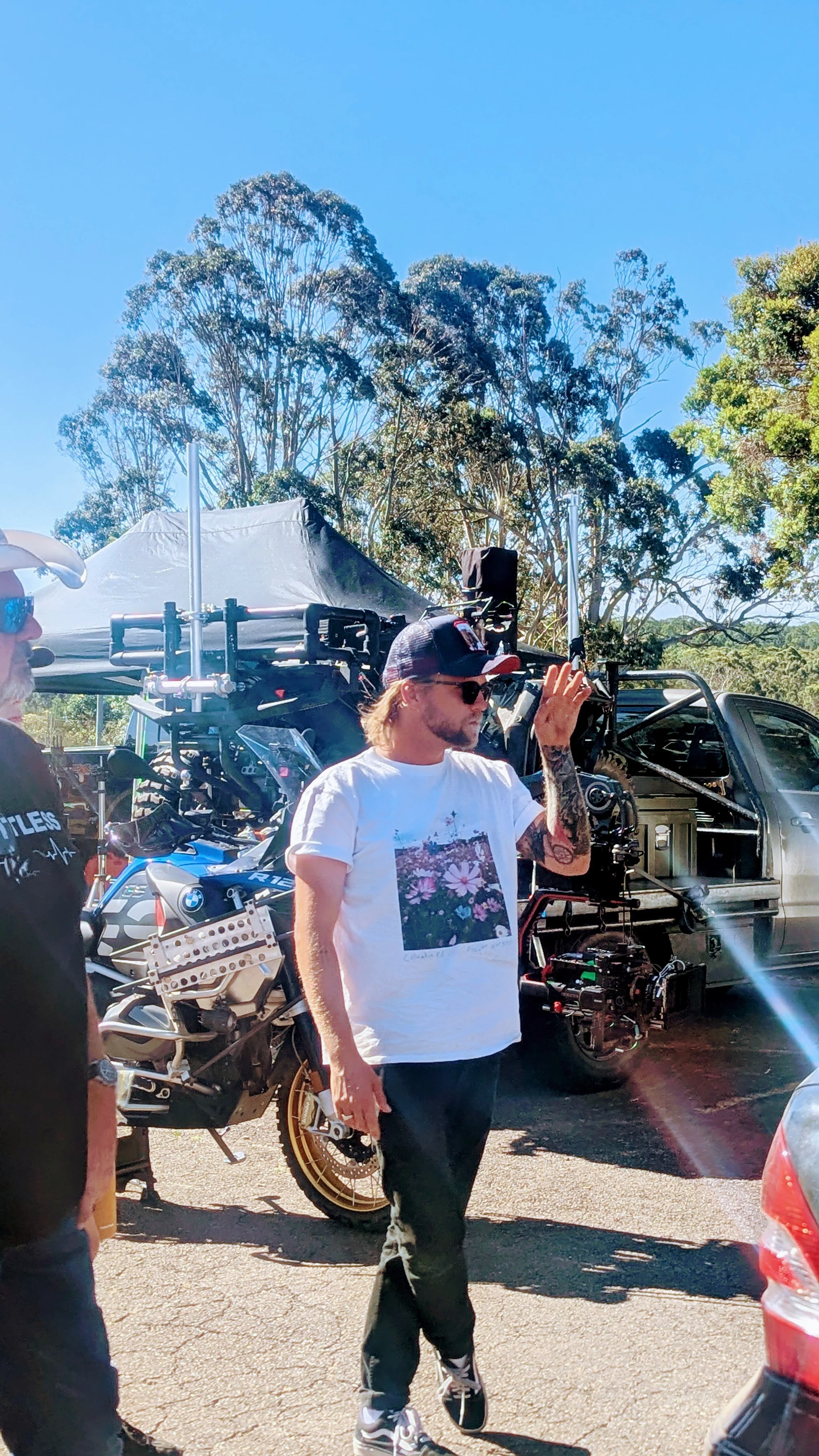 Man with tattoos and sunglasses waving next to a film camera and equipment, with a motorcycle and a truck in the background, outdoors on a sunny day.