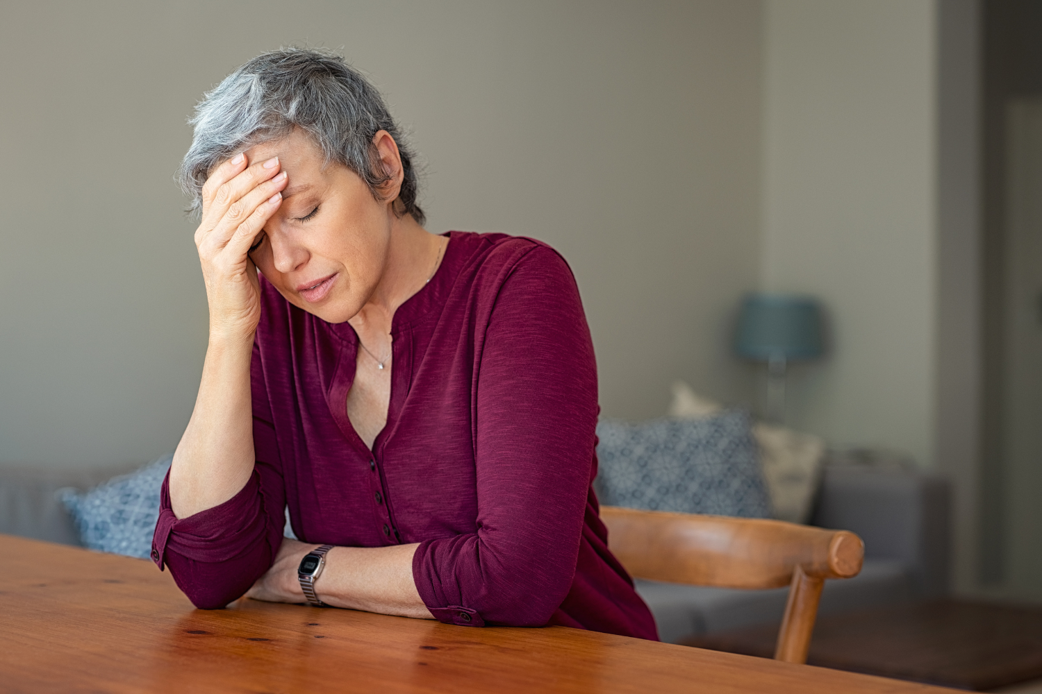 Vrouw zit aan tafel en houdt haar hoofd vast; ze oogt moe en overprikkeld.