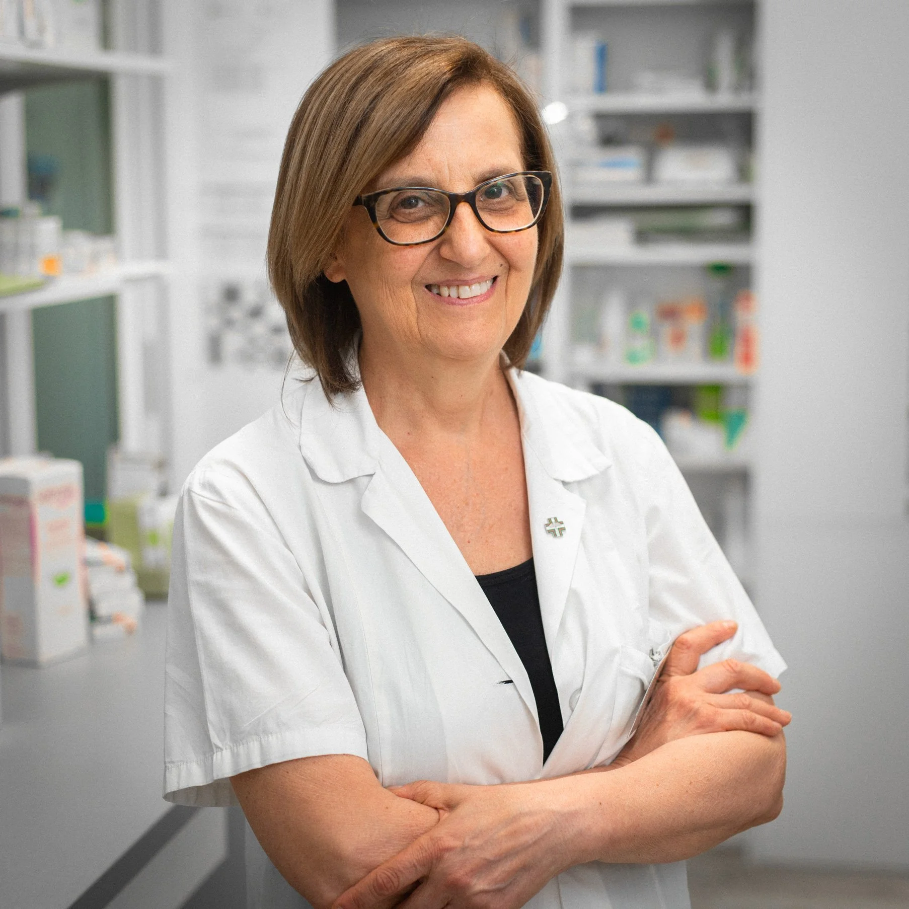 Una donna con capelli corti e occhiali indossa una giacca bianca da professionista e sorride, con le braccia incrociate, in un ambiente clinico o da laboratorio