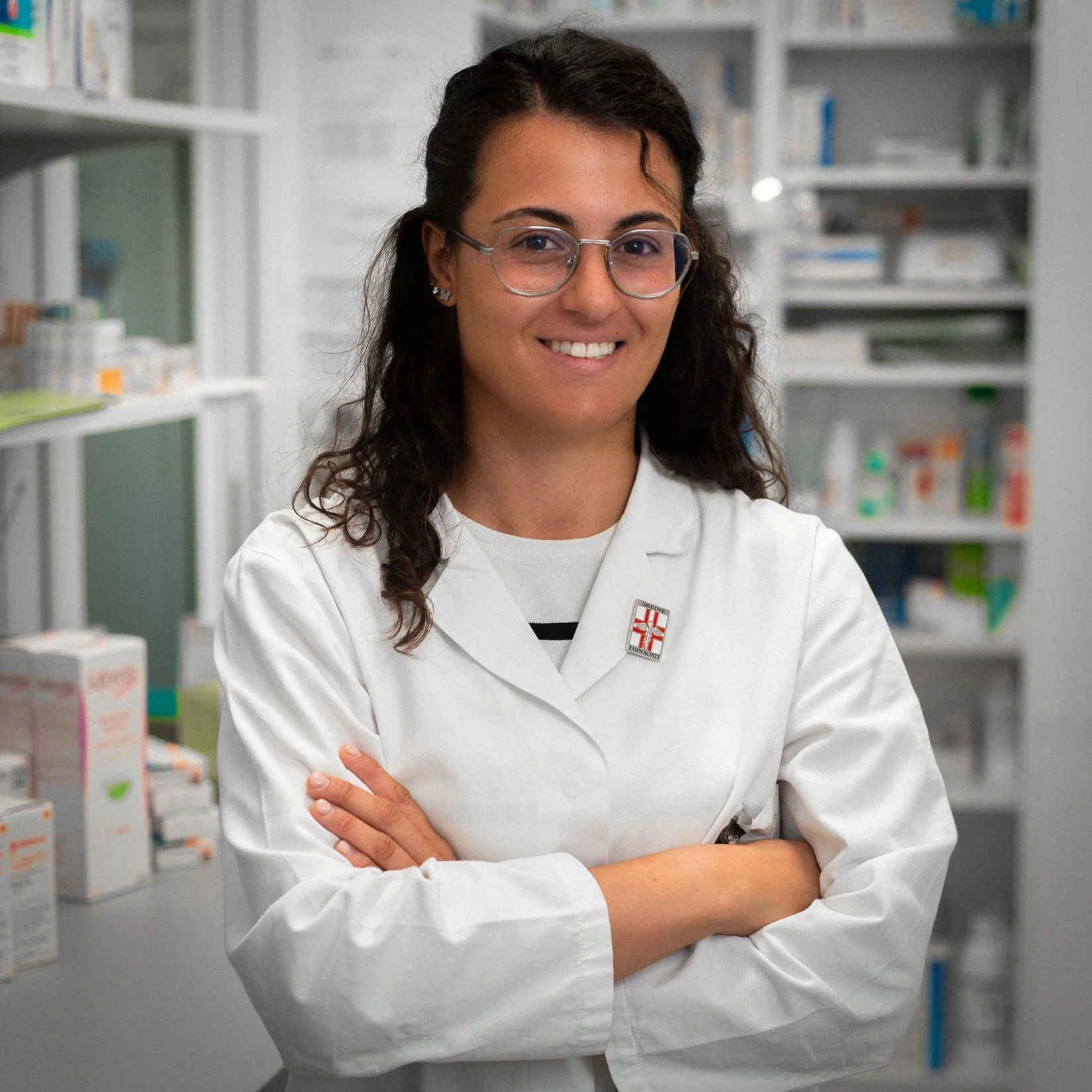 Una donna con occhiali, abbigliamento da laboratorio, sorride e si trova in una farmacia o laboratorio farmaceutico con scaffali pieni di confezioni di farmaci sullo sfondo.