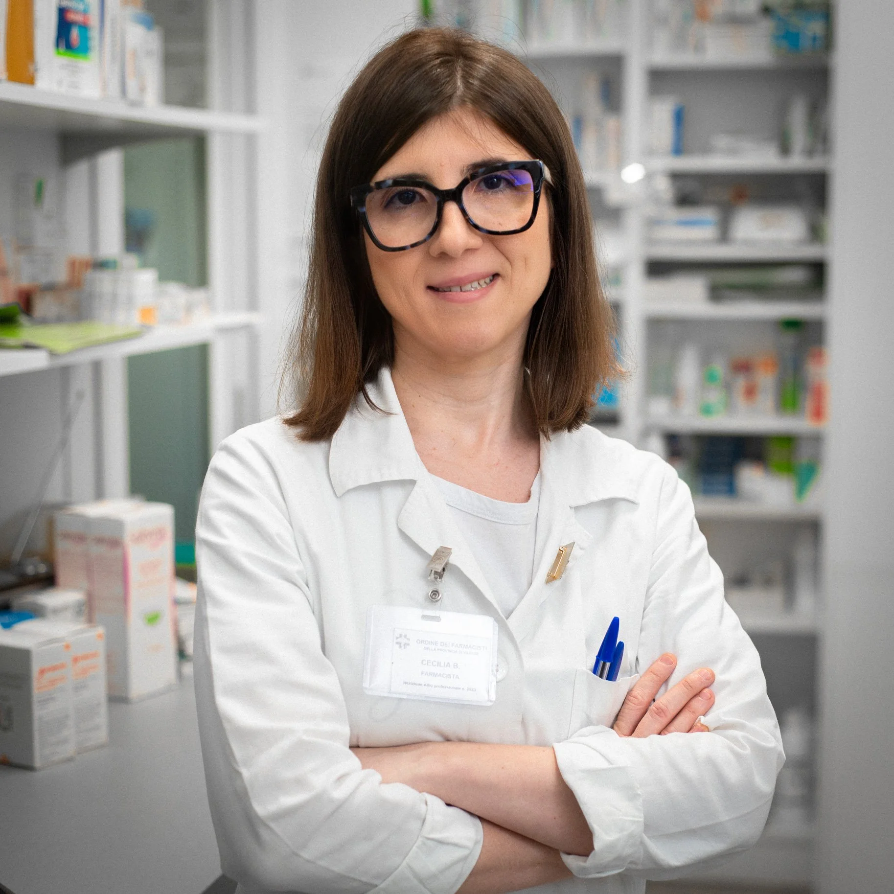 Donna farmacista con occhiali e camice bianco, in farmacia, con braccia incrociate, sorridente.