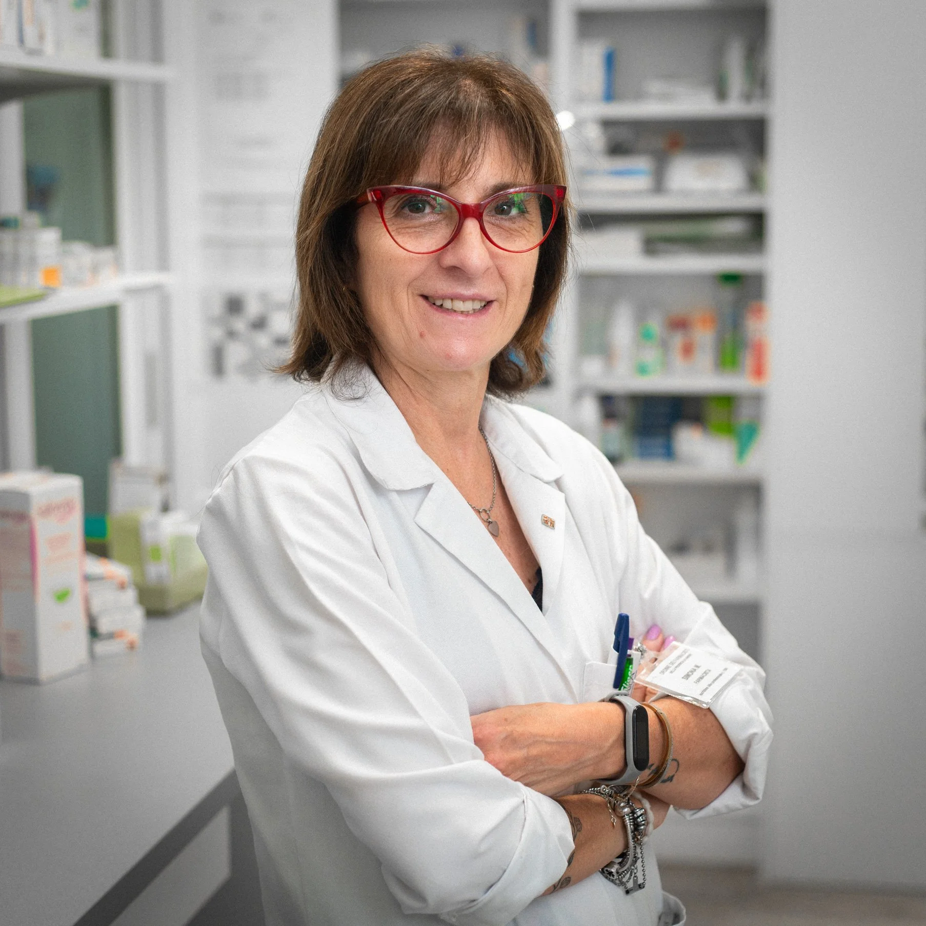 Una donna in camice bianco con occhiali rossi, in un ambiente di laboratorio o farmacia.