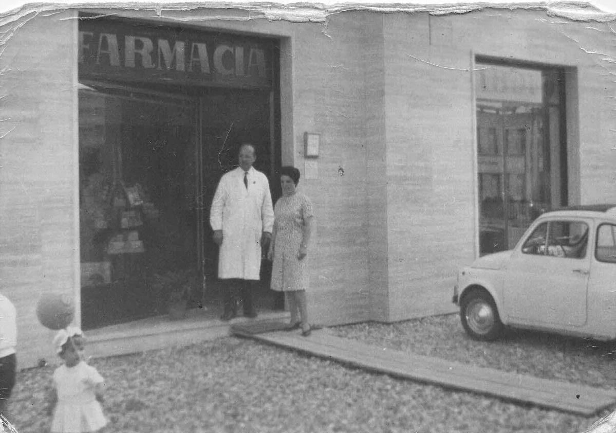 Una farmacia con due persone davanti, un uomo vestito da medico e una donna, con un'auto d'epoca parcheggiata vicino.