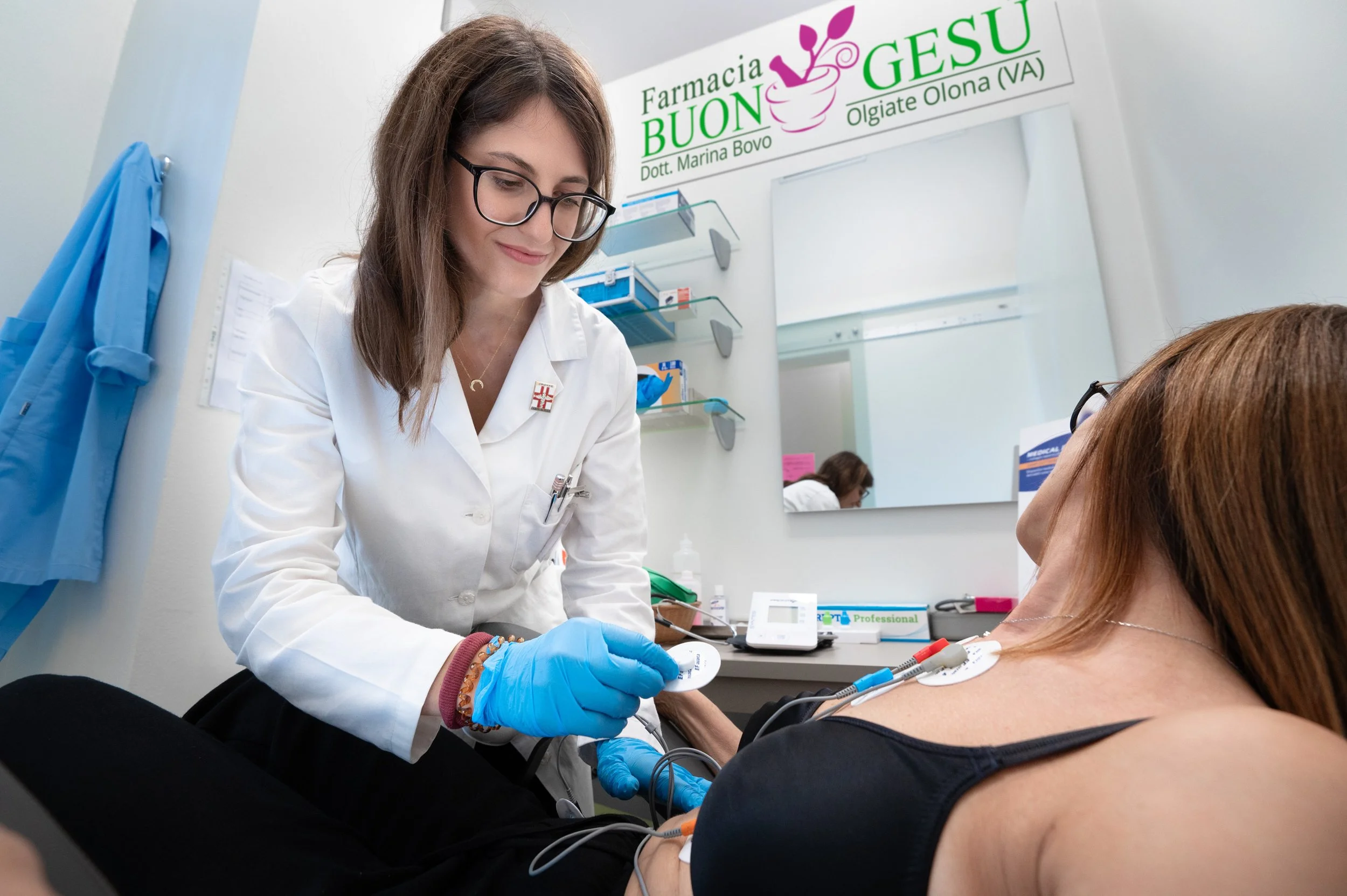 Una donna con occhiali e camice bianco che preleva sangue da una donna sdraiata su un letto di ospedale, appare un ambiente clinico.