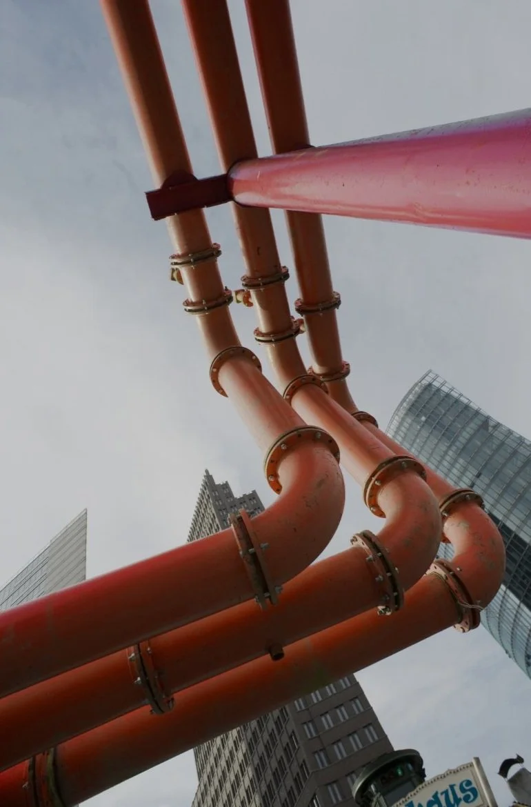 Close-up of orange industrial pipes against city buildings and a cloudy sky.