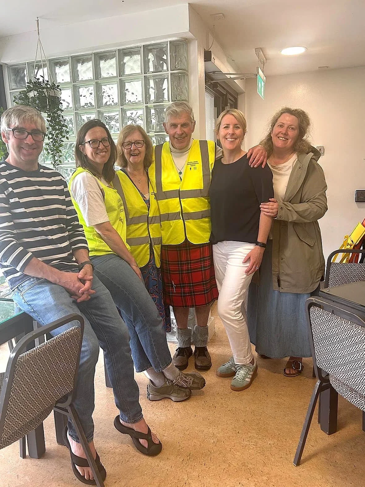 Group of LLEG members standing indoors, smiling, with a glass brick wall and potted plant in the background.
