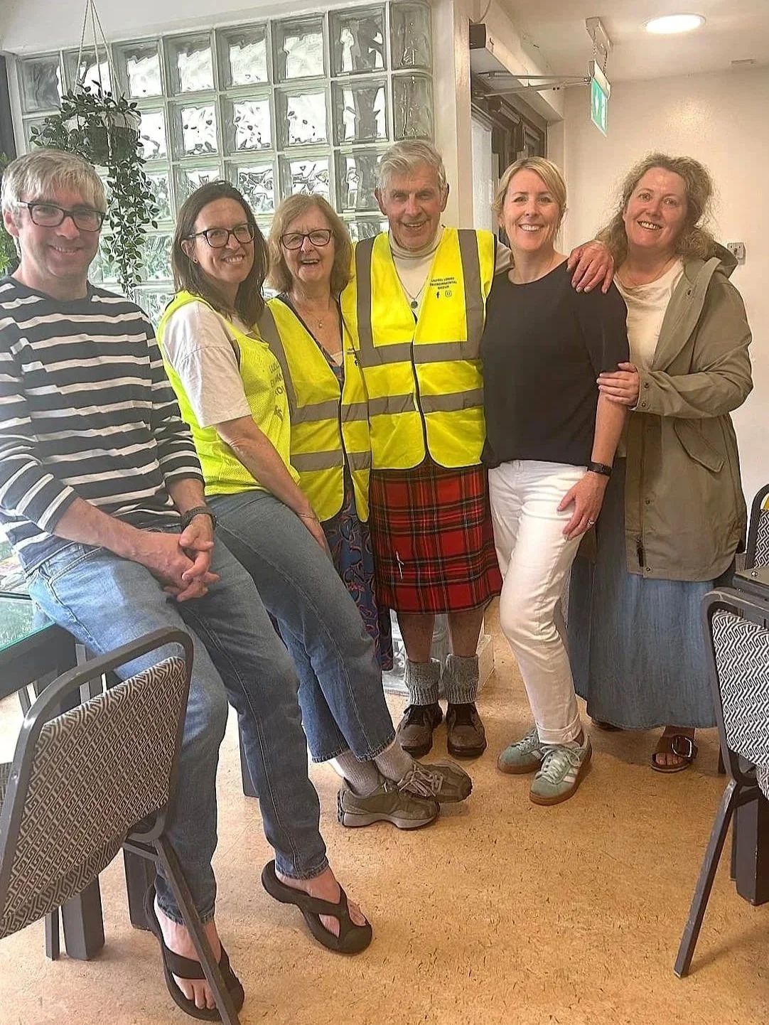 Group of six LLEG members smiling, three of whom are wearing yellow safety vests. The group is standing inside a room with a glass block wall and potted plant in the background.