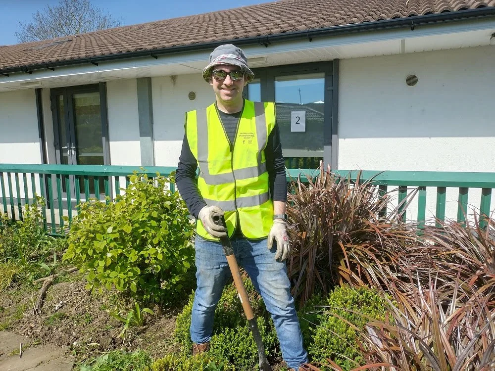 Member of LLEG group wearing sunglasses, and a yellow safety vest stands outdoors in a garden, holding a shovel, smiling at the camera.
