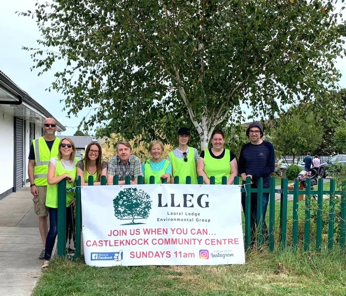 Group of LLEG members standing behind a fence with a banner, wearing bright yellow vests, in front of a castleknock community center, promoting community environmental activity.