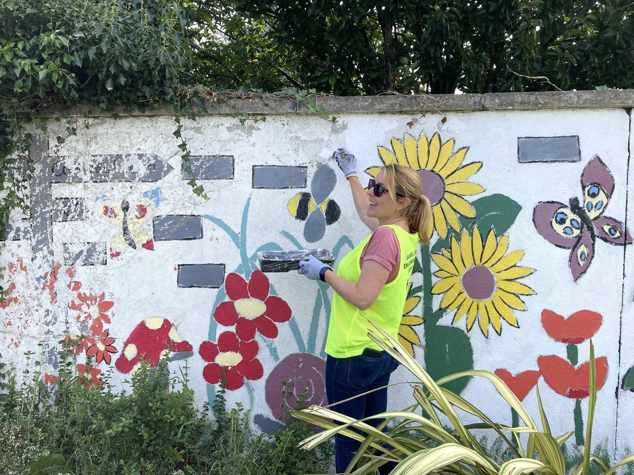Woman, member of Laurel Lodge Environmental Group painting a colorful mural of flowers and butterflies on a white wall using brushes.