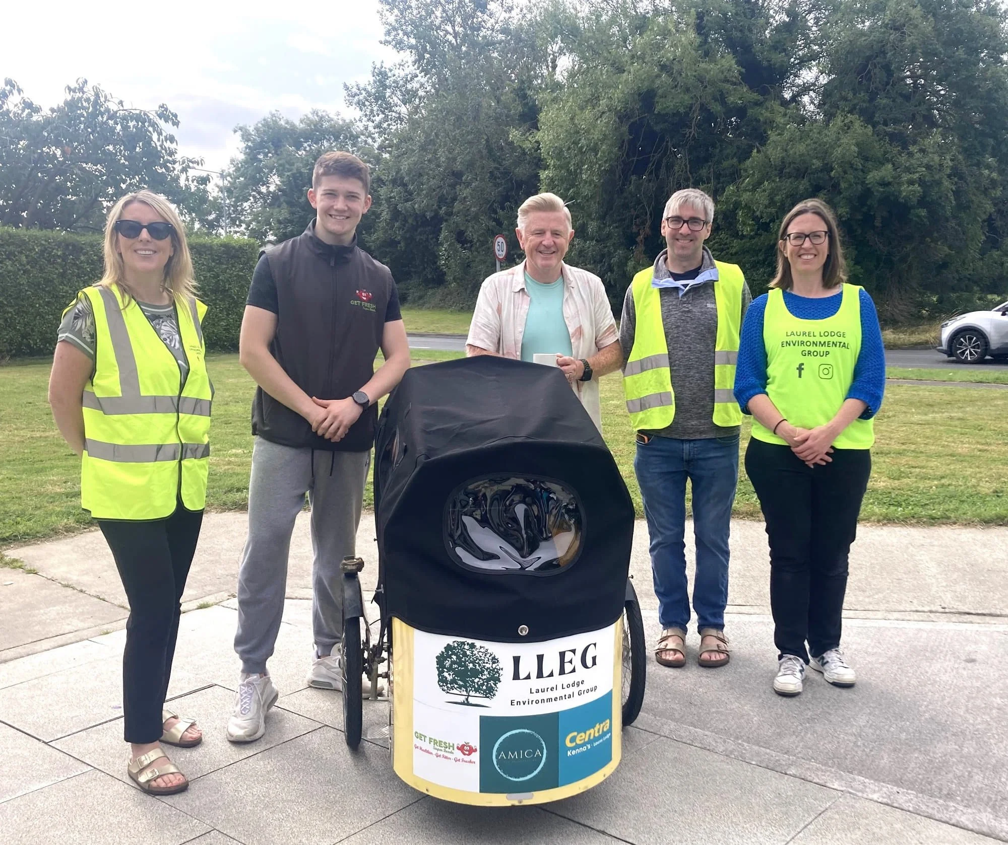 Group of LLEG sponsors, with a baby stroller covered with a sign reading 'LLeg Laurel Lodge Environmental Group' and various logos, in front of green bushes and trees.