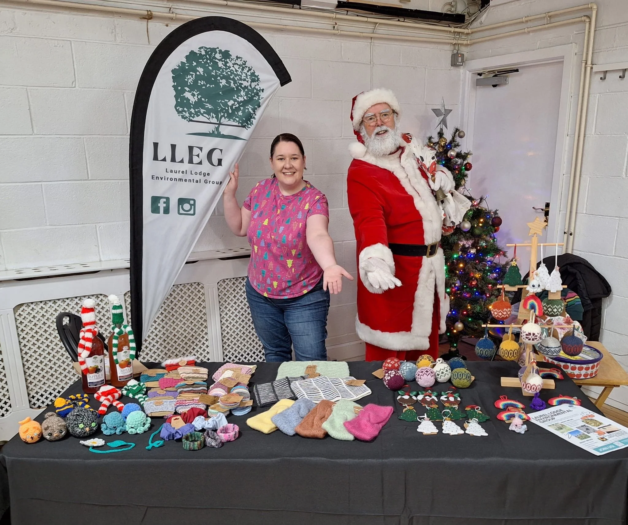 A woman smiling at a holiday craft table with a man dressed as Santa Claus beside her. The table displays various handmade holiday craftsA Christmas tree decorated with lights and ornaments is in the background, along with a vertical banner for Laurel Lodge Environmental Group.