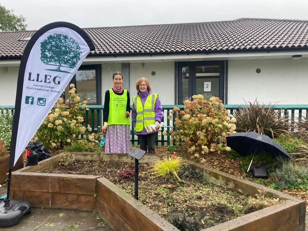 Two women holding a planting tool and wearing biosecurity vests stand in front of a raised garden bed outside castleknock community center. A black umbrella is on the ground nearby, and there is a flag with the Laurel Lodge Environmental Group logo.