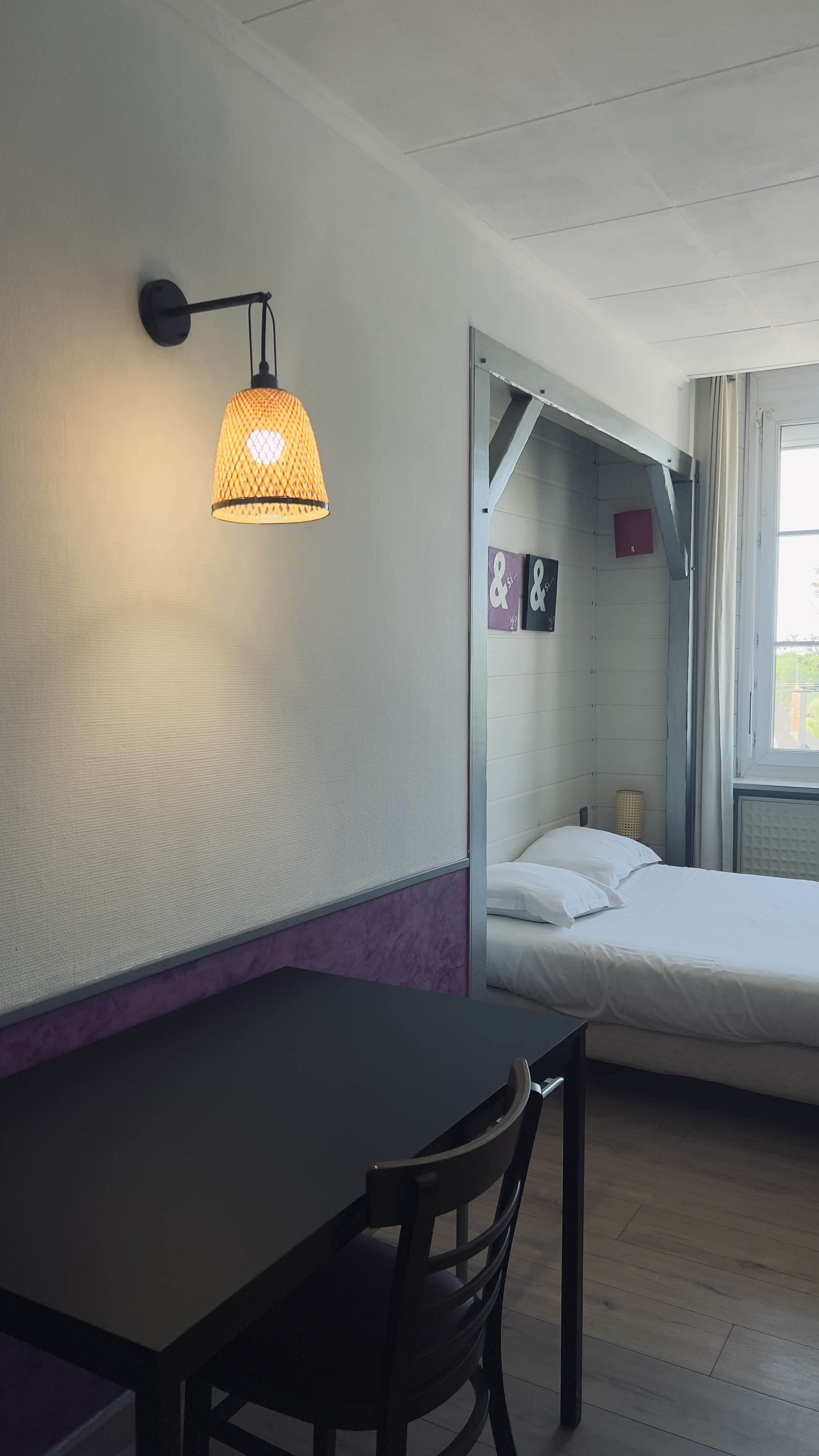Chambre avec un lit, une table noire, une chaise, une lampe murale allumée, et une fenêtre.