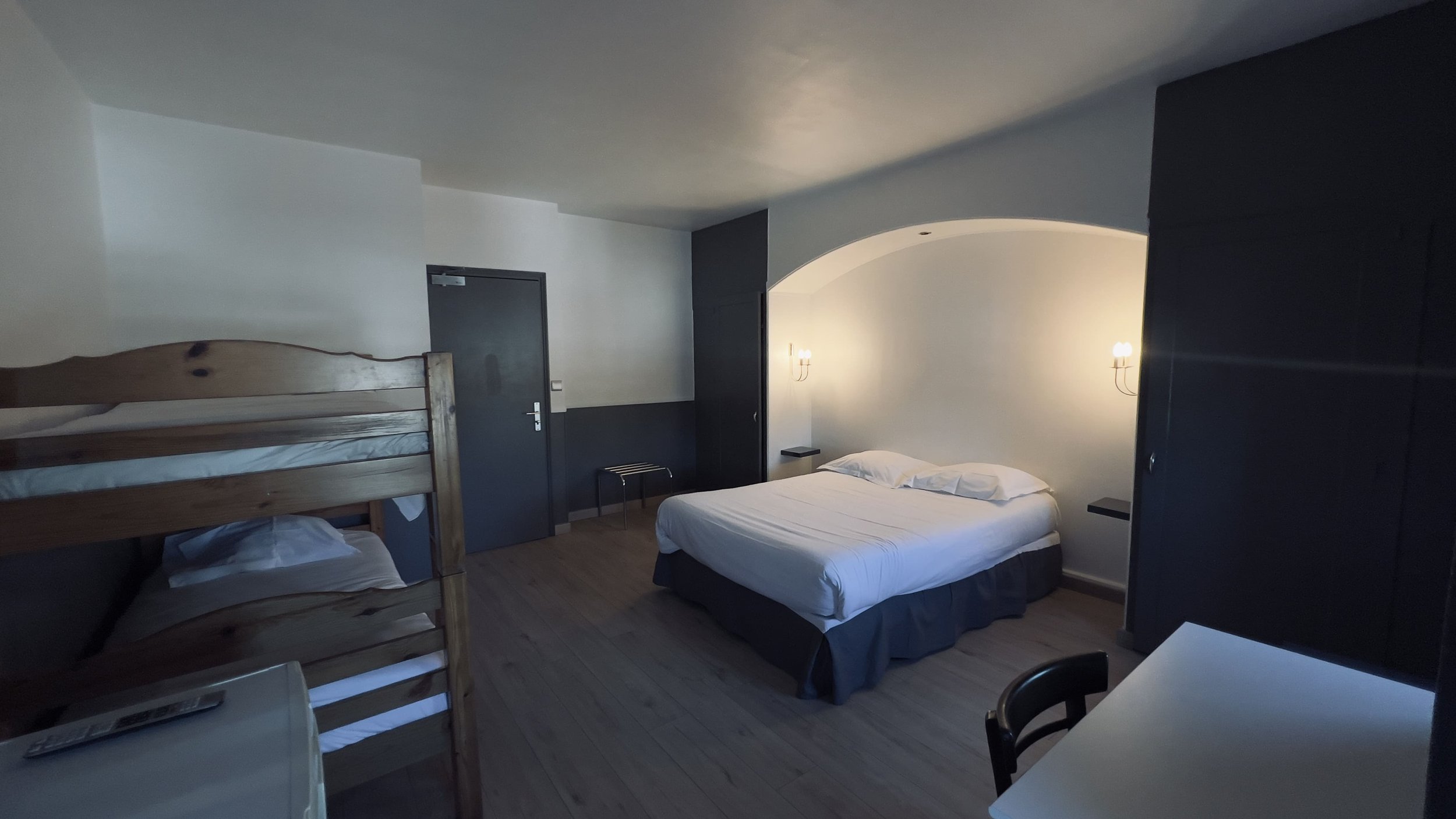 Chambre d'hôtel avec lit double, lits superposés, table, et chaises, décor minimaliste.