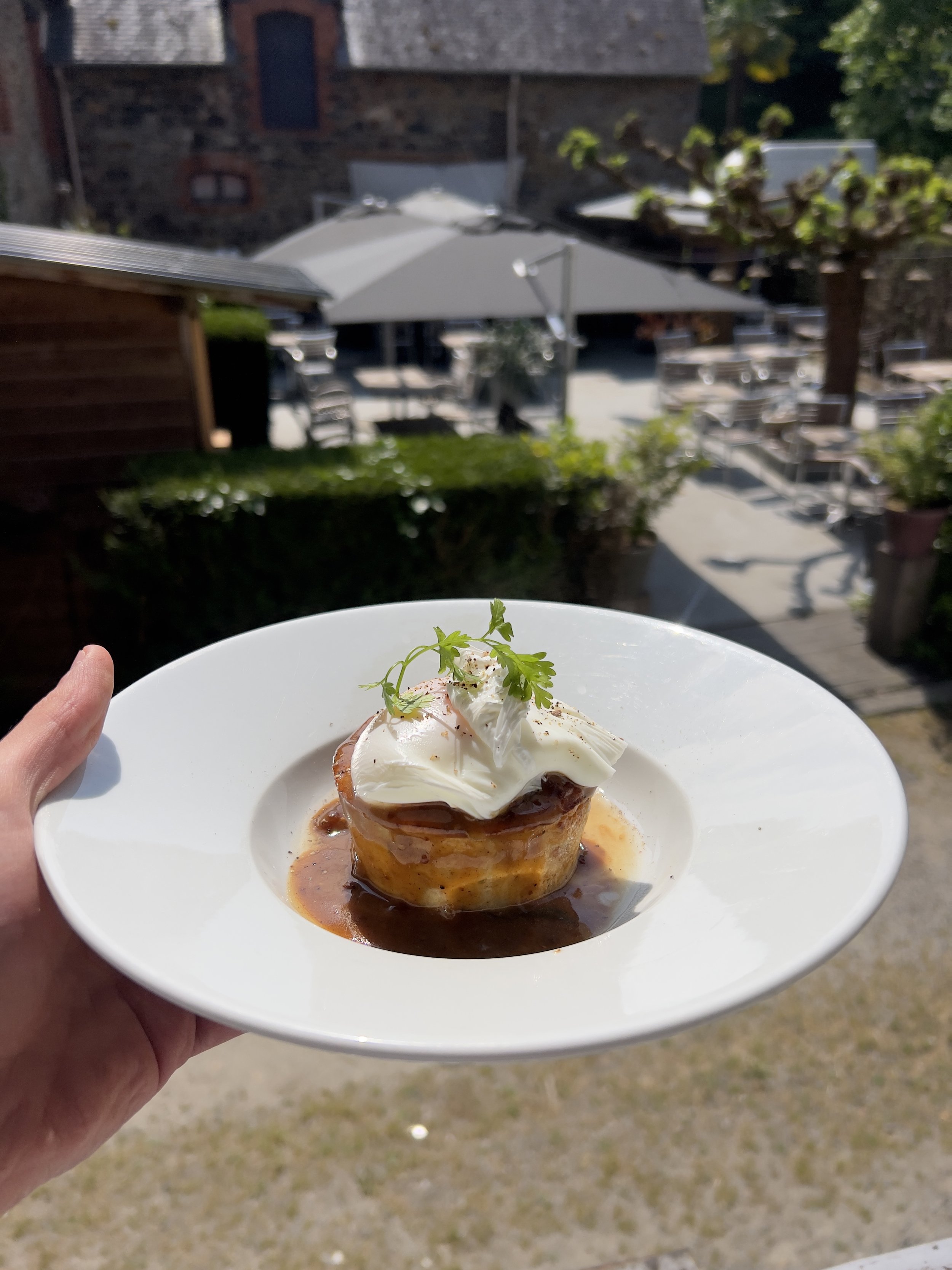 Main tenant une assiette avec un plat gourmet à base de pommes de terre et œuf poché, garni de sauce et herbes, en extérieur.