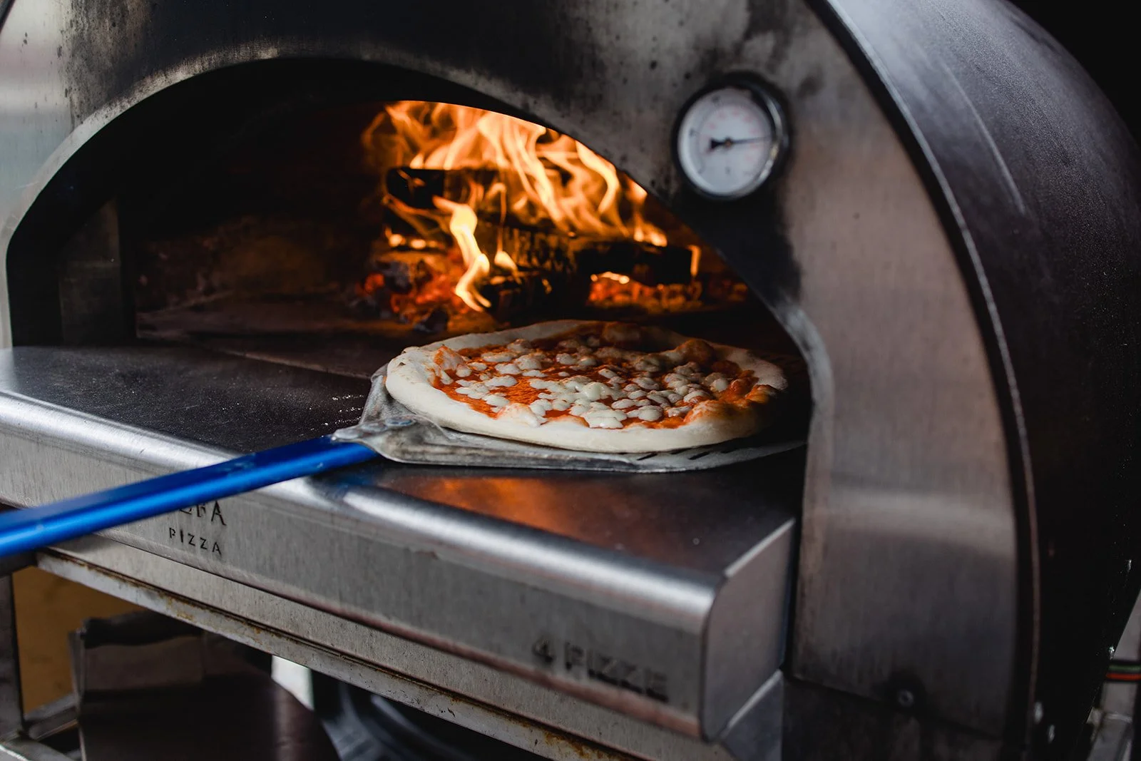 Eine Pizza wird in einem Holzofen gebacken, eine Flamme ist im Hintergrund sichtbar.