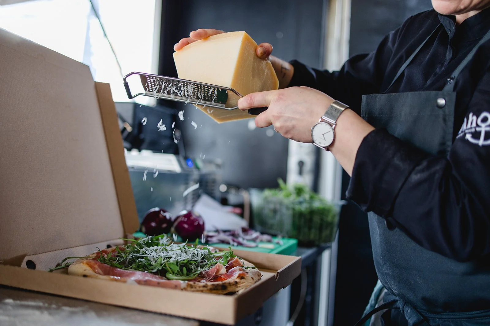 Koch reibt Käse über eine Pizza mit Rucola und Schinken in einem Pizzakarton.