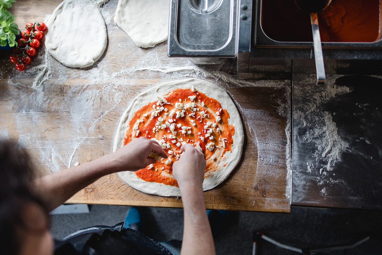 Ein Mensch bereitet eine Pizza zu. Der Pizzateig wird mit Tomatensauce und Käse belegt. Auf dem Tisch liegen weitere Teigstücke, Tomaten und frisches Basilikum. Eine Küchentheke mit Metallbehältern ist sichtbar.
