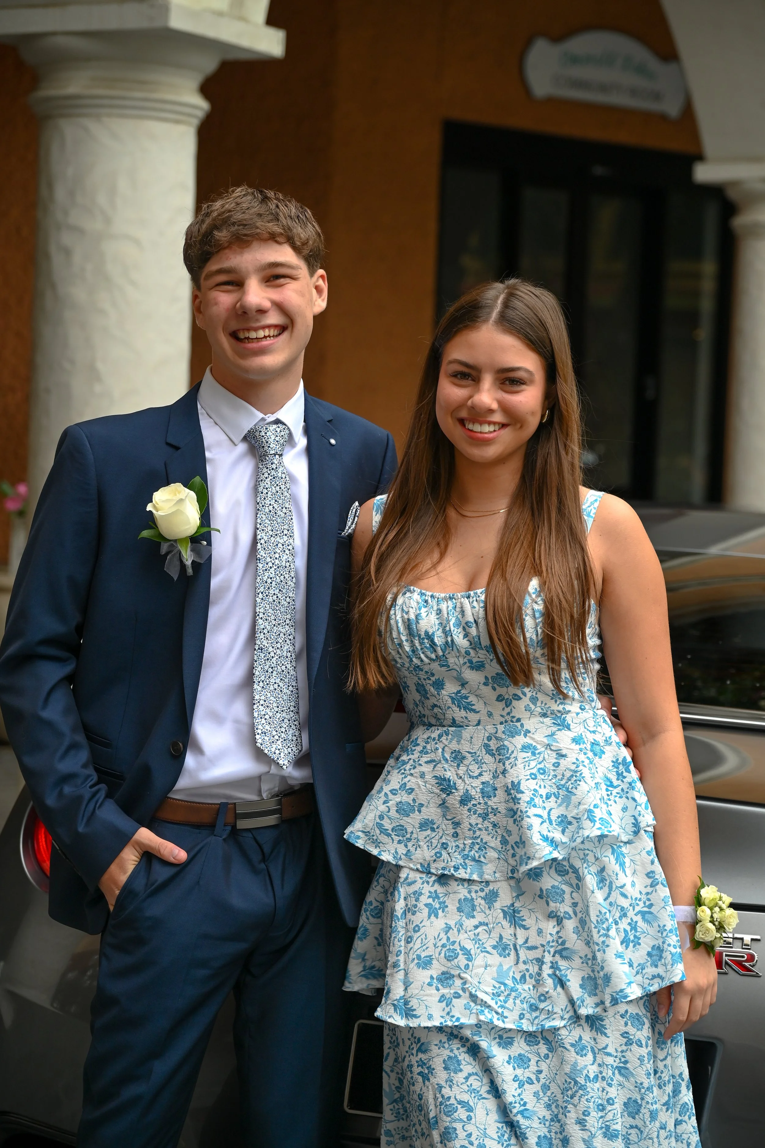 A young man in a navy blue suit with a white shirt and patterned tie, and a young woman in a blue and white floral dress, smiling and standing outdoors in front of a car, at a celebration or special event.