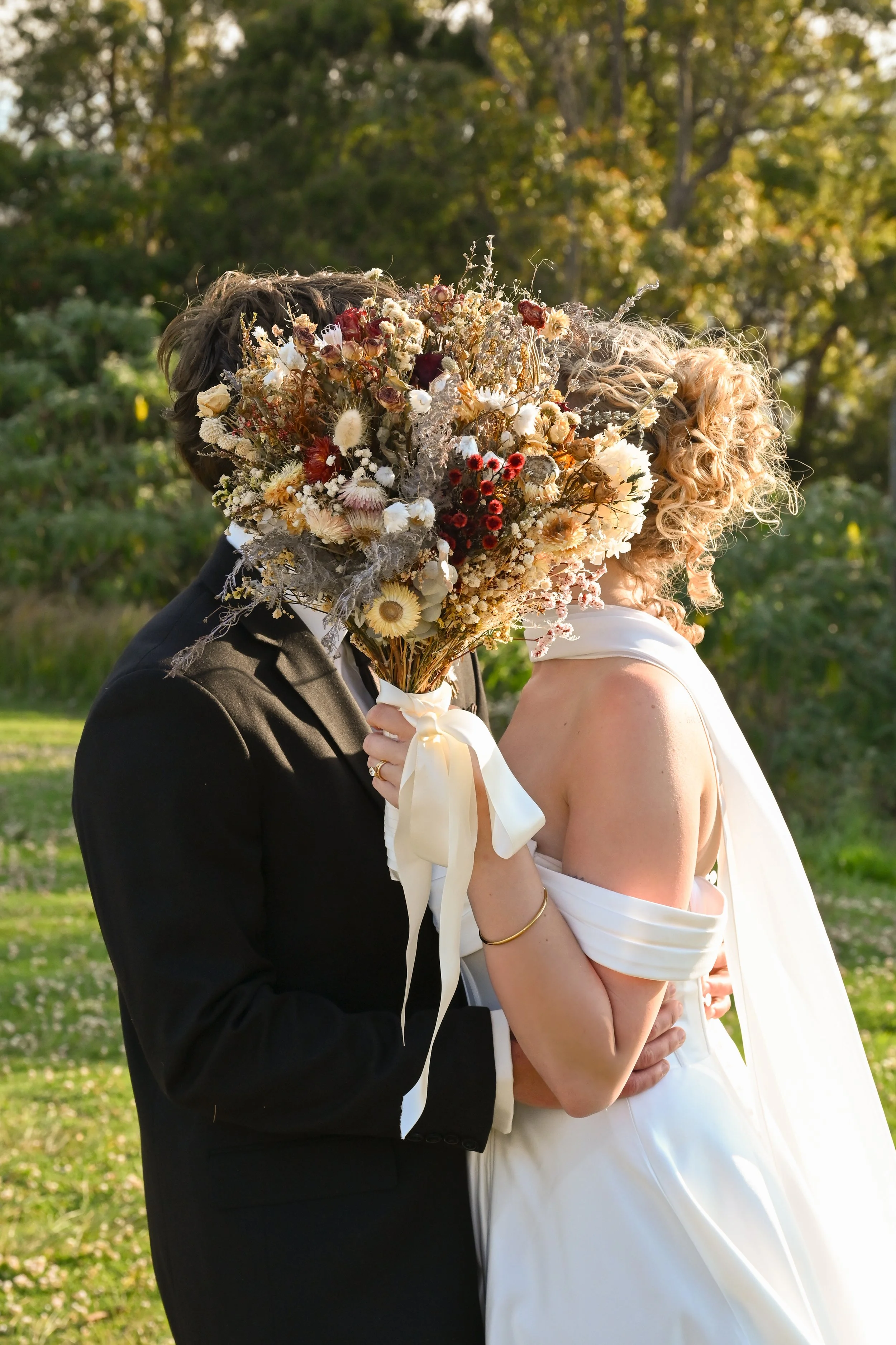 A couple on their wedding day, holding a large bouquet of dried flowers in front of their faces, outdoors with green trees and grass in the background.