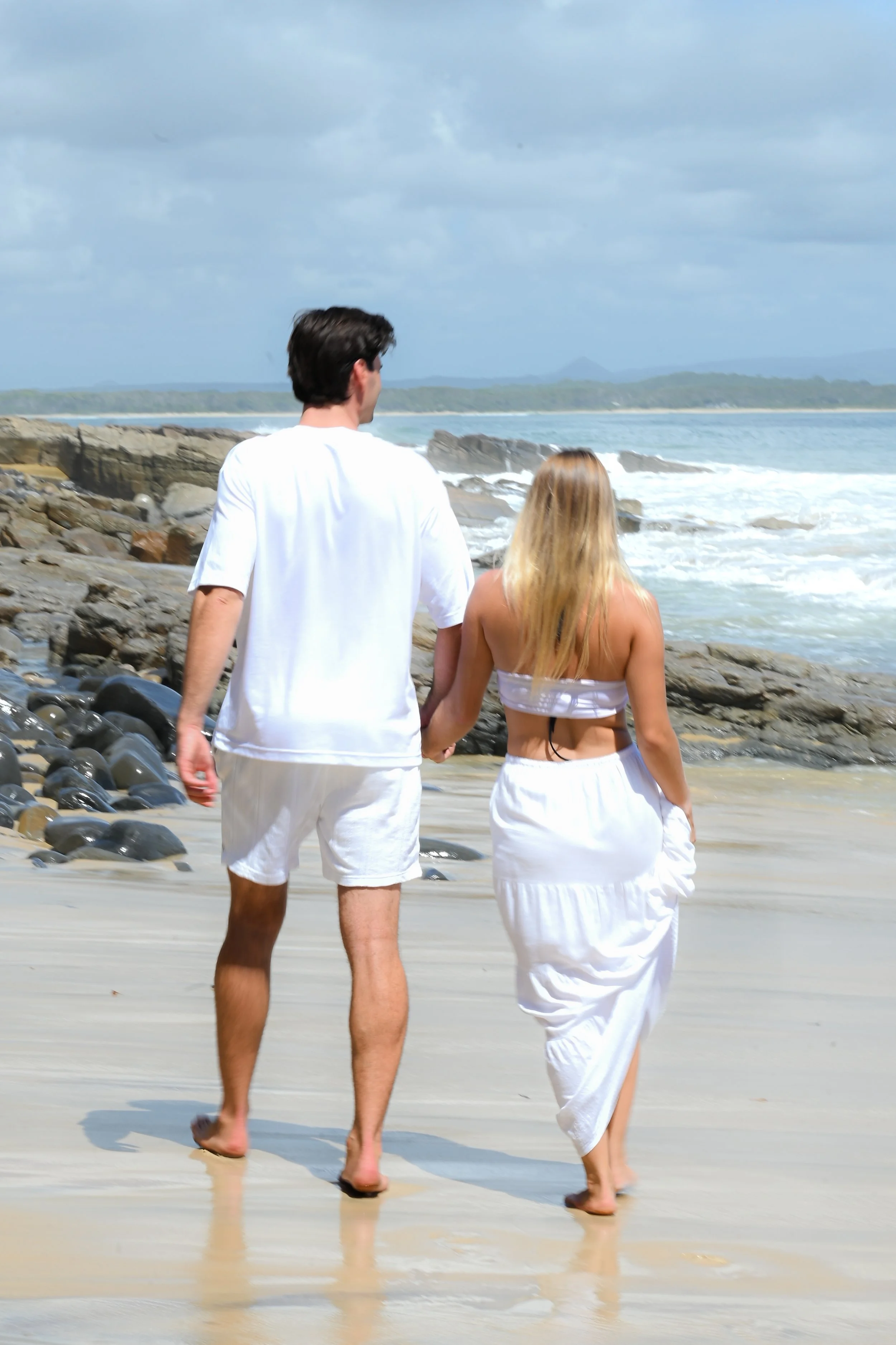 A young couple walking hand-in-hand on a beach with rocks and ocean waves, wearing white beach clothing.