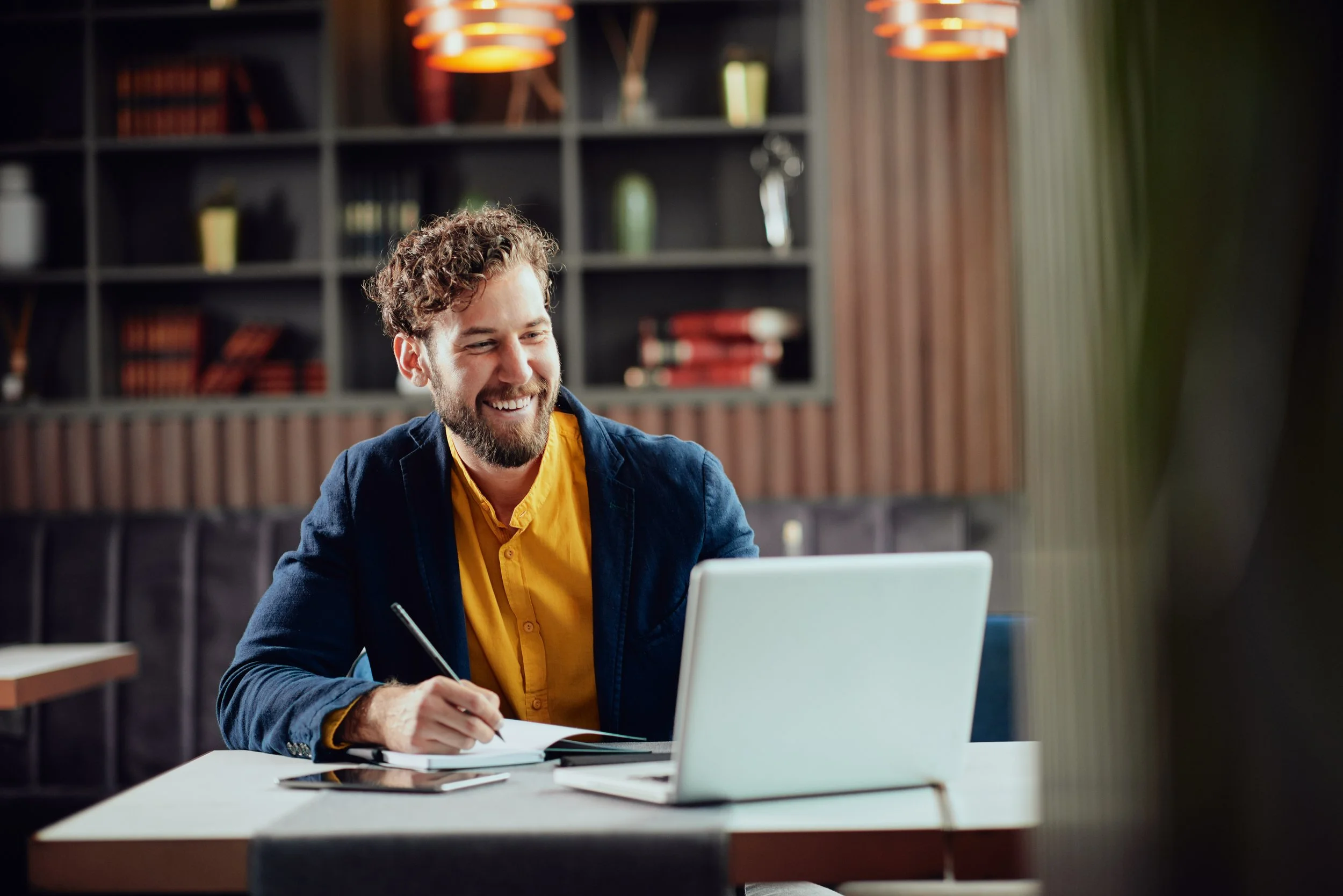 Man working on a laptop in a modern office, writing in a notebook, smiling, bookshelves in the background.