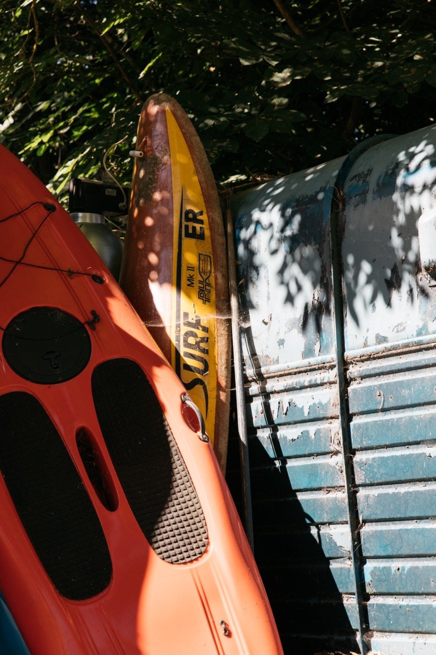 Orange kayak and yellow surfboard leaning against a blue metal surface surrounded by shadows from nearby foliage.