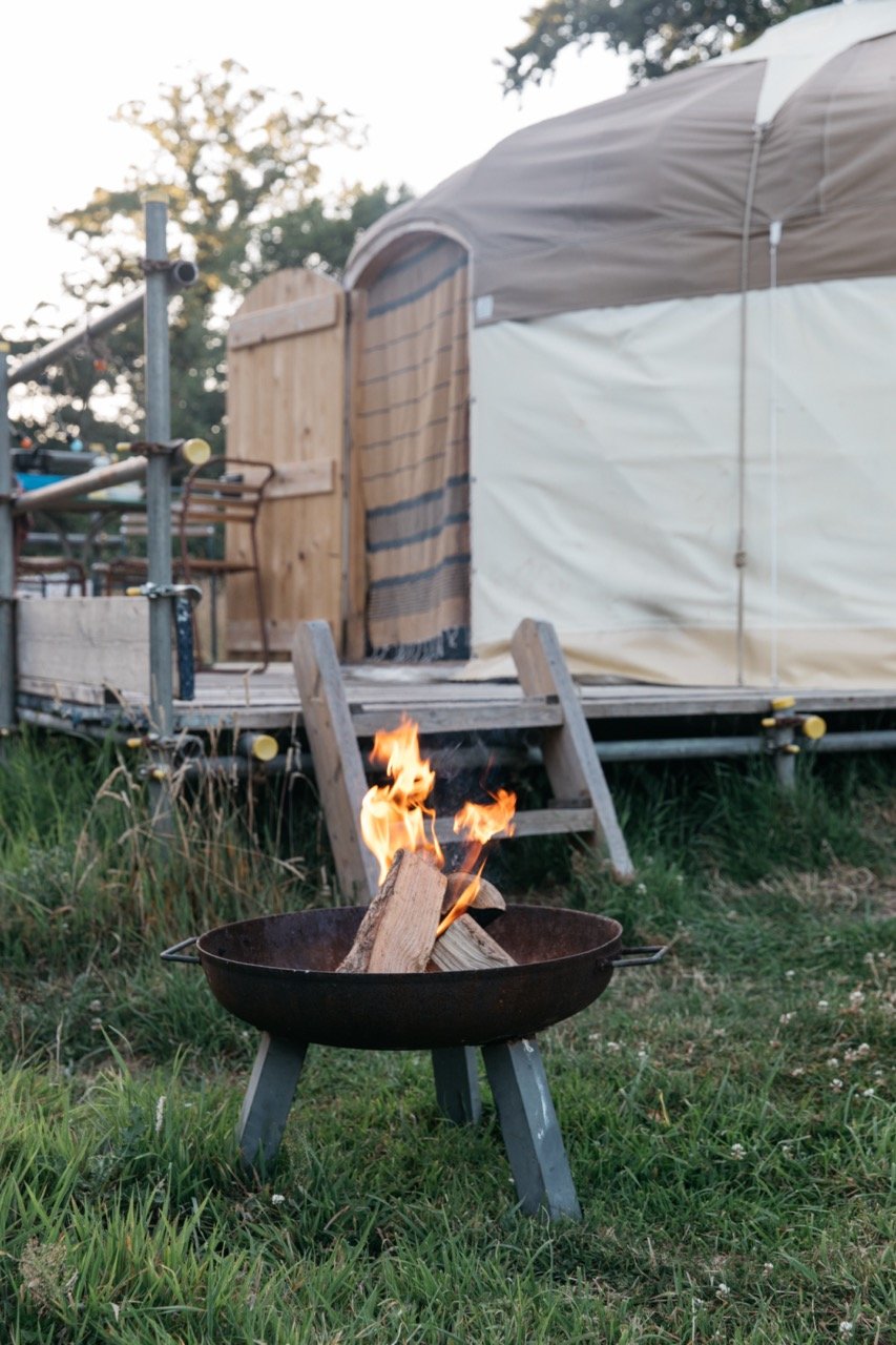 Outdoor fire pit with burning logs in front of a yurt tent on grassy area.
