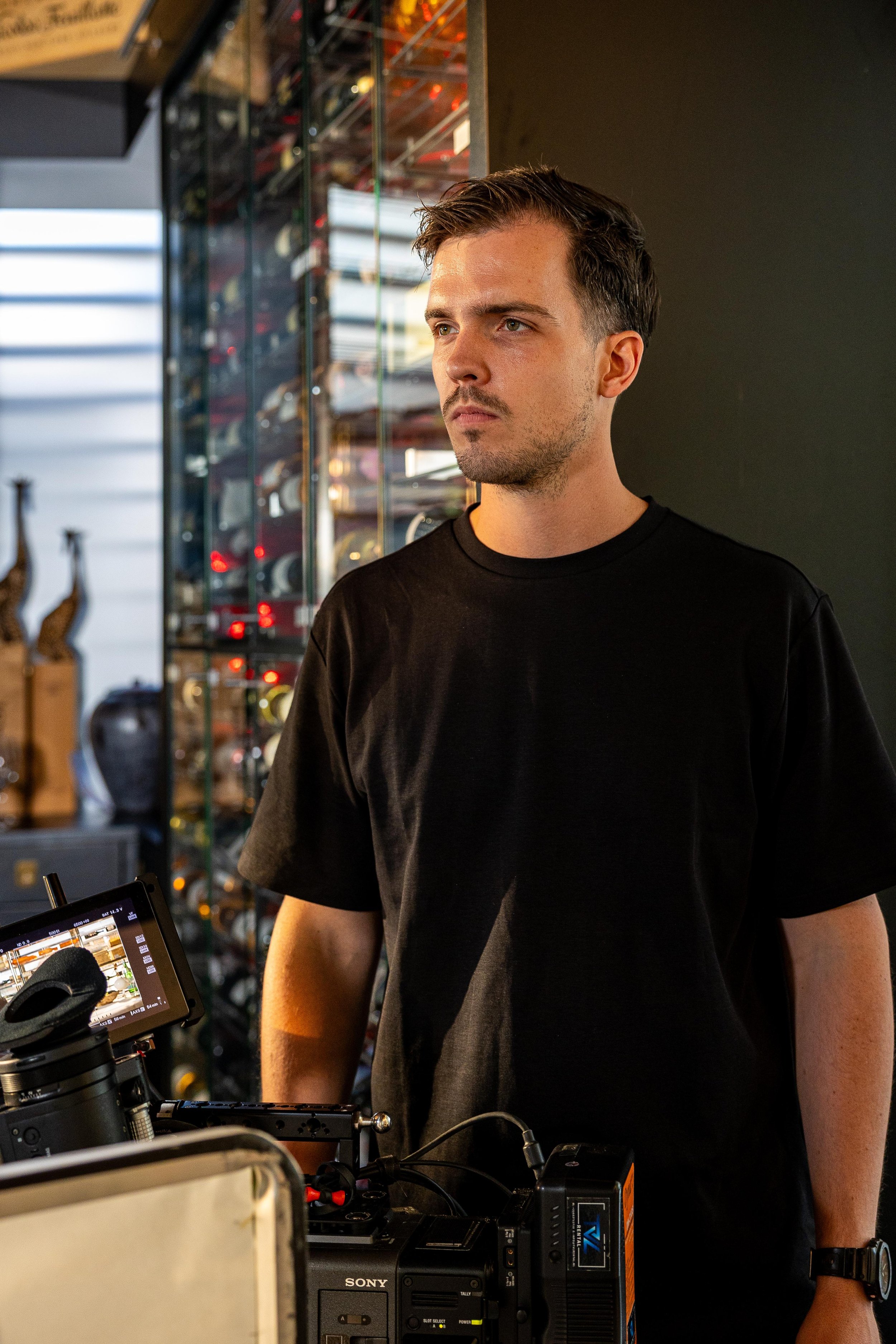 Een man met kort bruin haar en een zwarte T-shirt staat naast fotobewerking apparatuur, mogelijk op een filmset of in een studio.