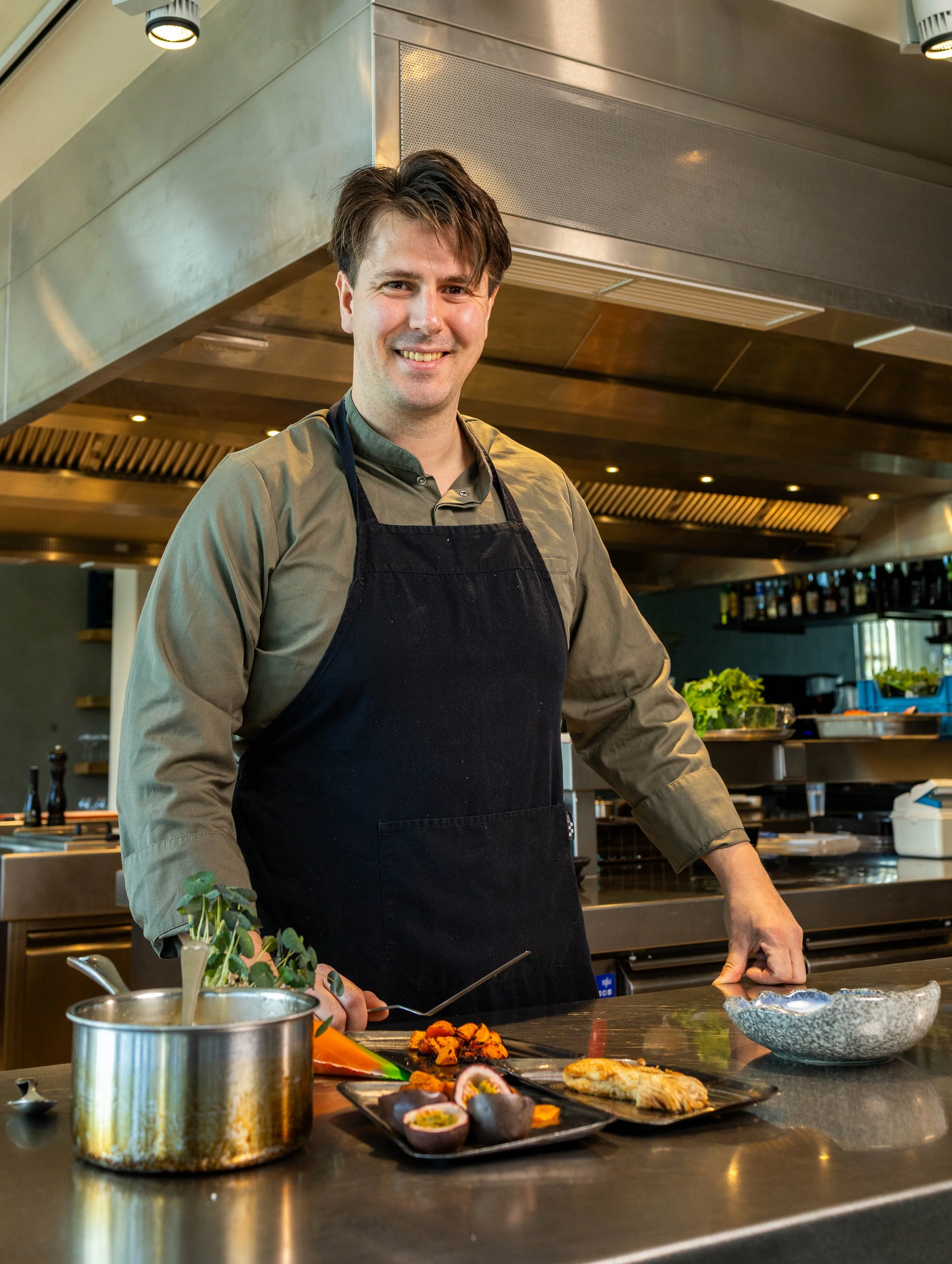 Man in een keuken, glimlachend, gekleed in een grijze blouse en een zwart schort, bereidt voedsel met diverse ingrediënten op het aanrecht.