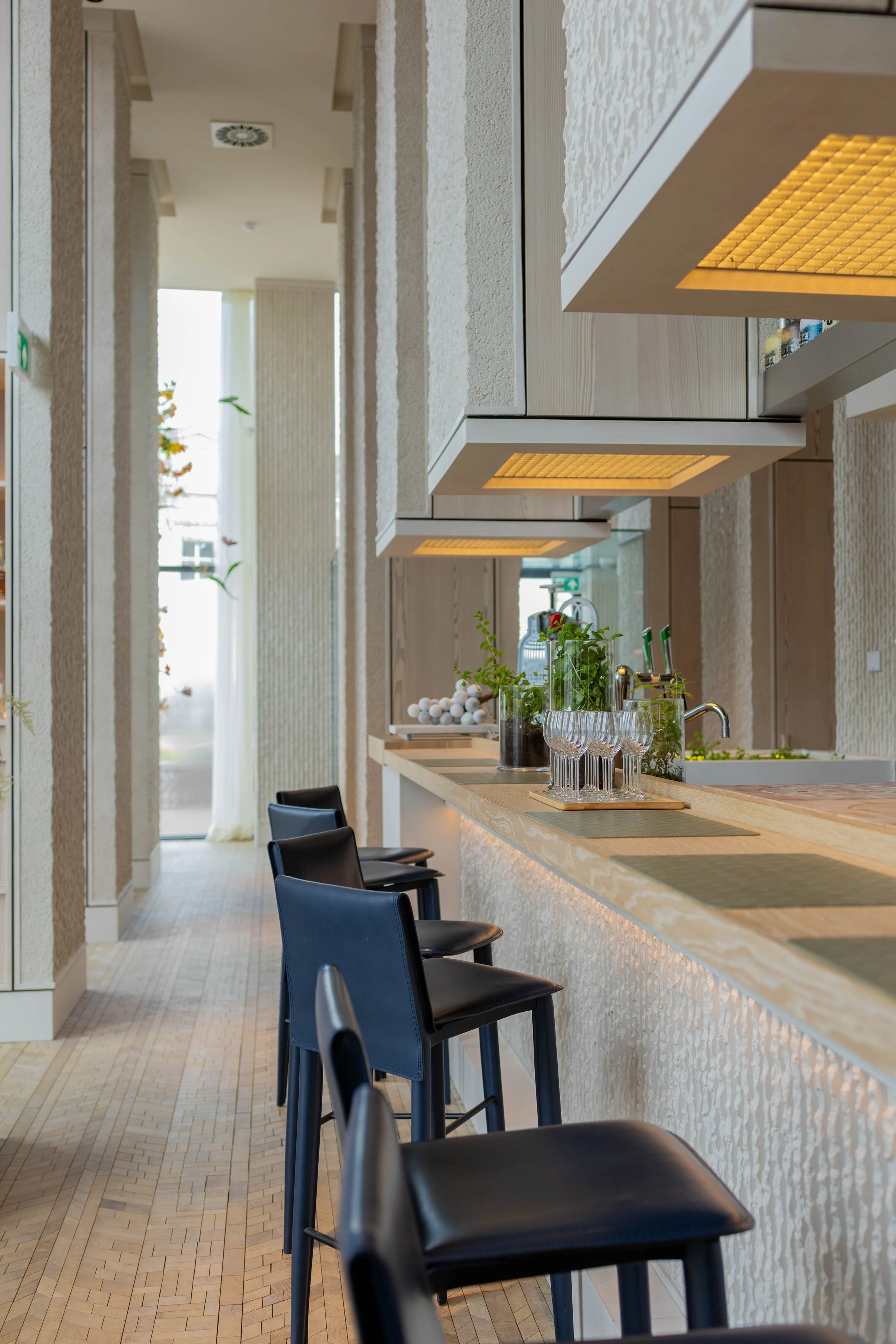 Een keukeneiland met vier zwarte barkrukken, decoratief planten, glazen en keukengerei, boven een bar met bergruimte, binnen een lichte, moderne keuken met grote ramen en witte muurafwerking.