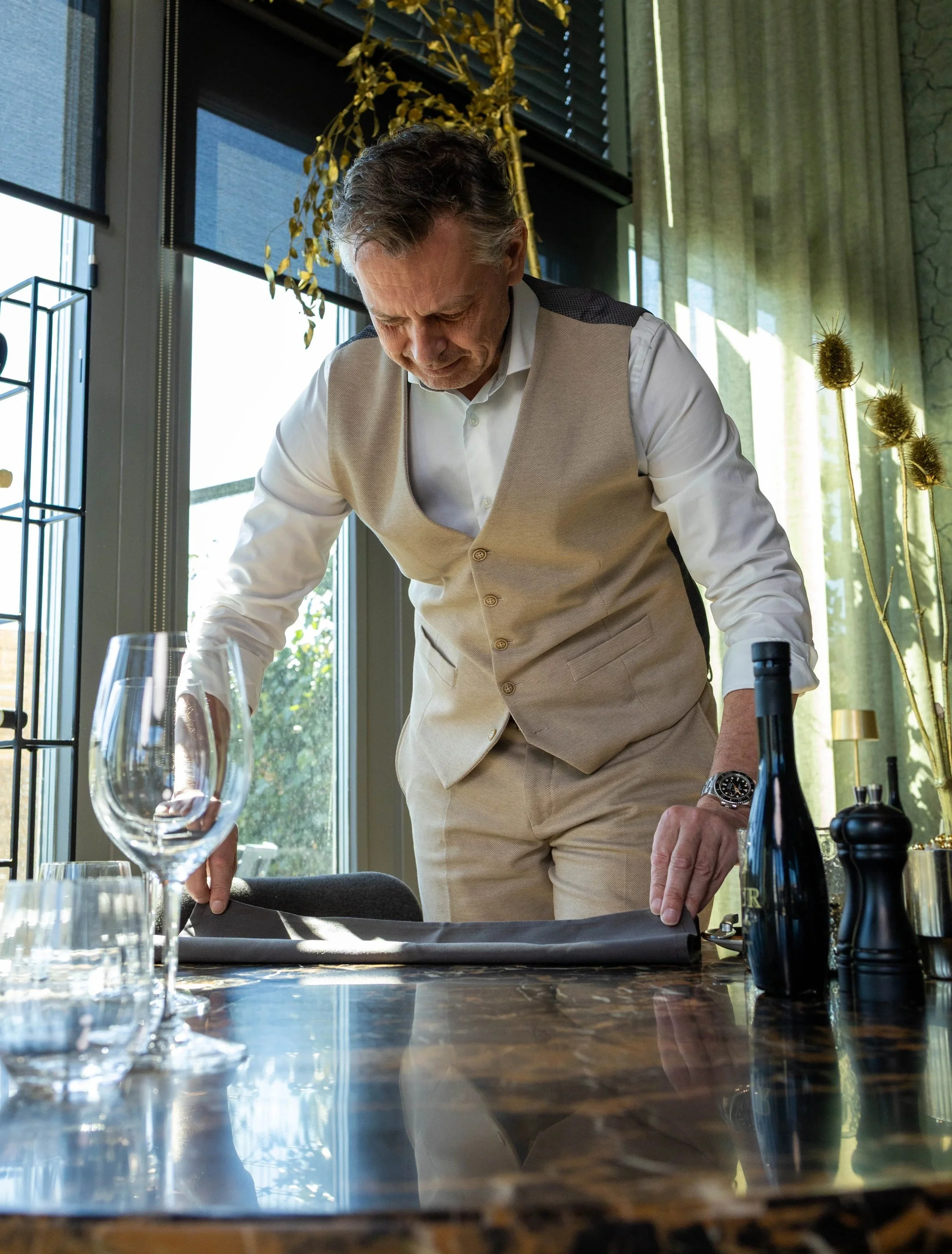 Man in een beige vest en wit overhemd die zich voorbereidt om te dineren in een restaurant met grote ramen en groene planten.