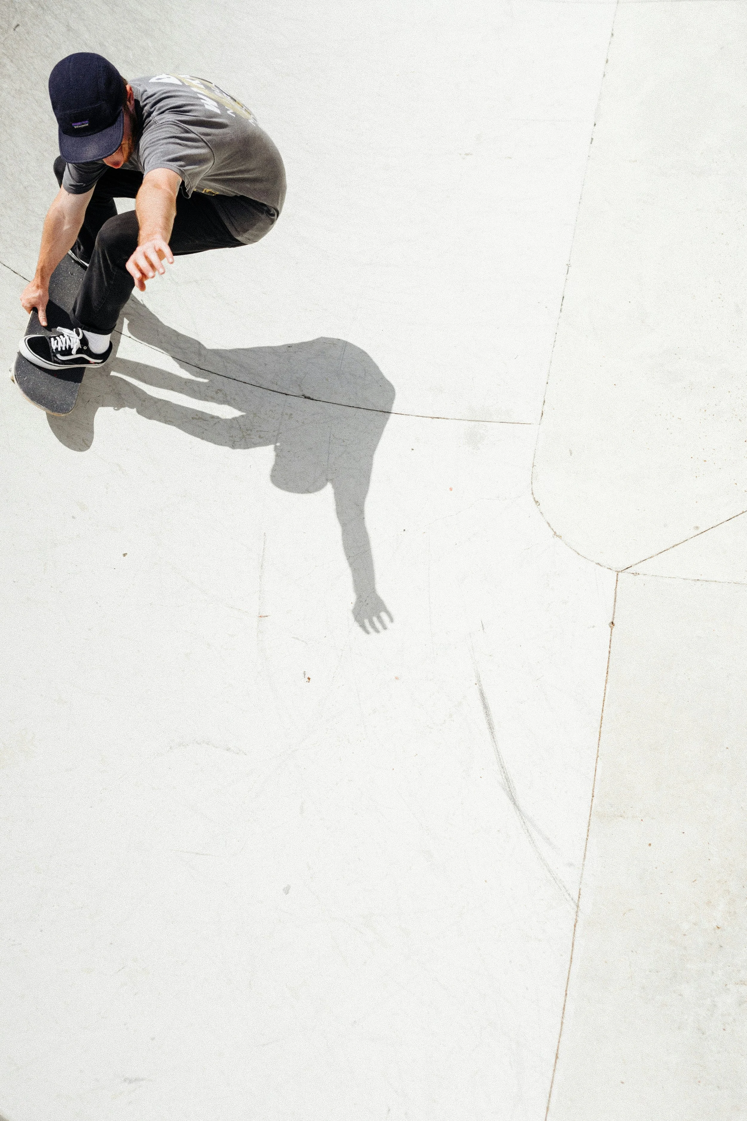 A person wearing a gray shirt, black pants, and a black cap skateboarding on a light-colored concrete surface, casting a shadow.