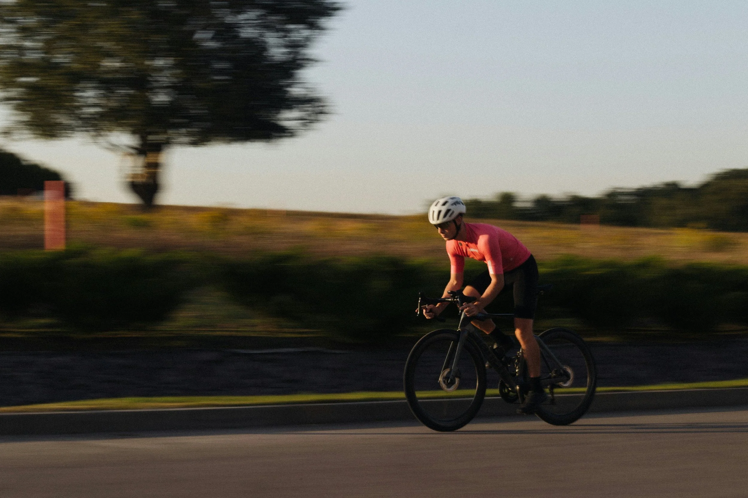 A person wearing a pink shirt, black shorts, and a white helmet riding a black bicycle on a road during sunset.