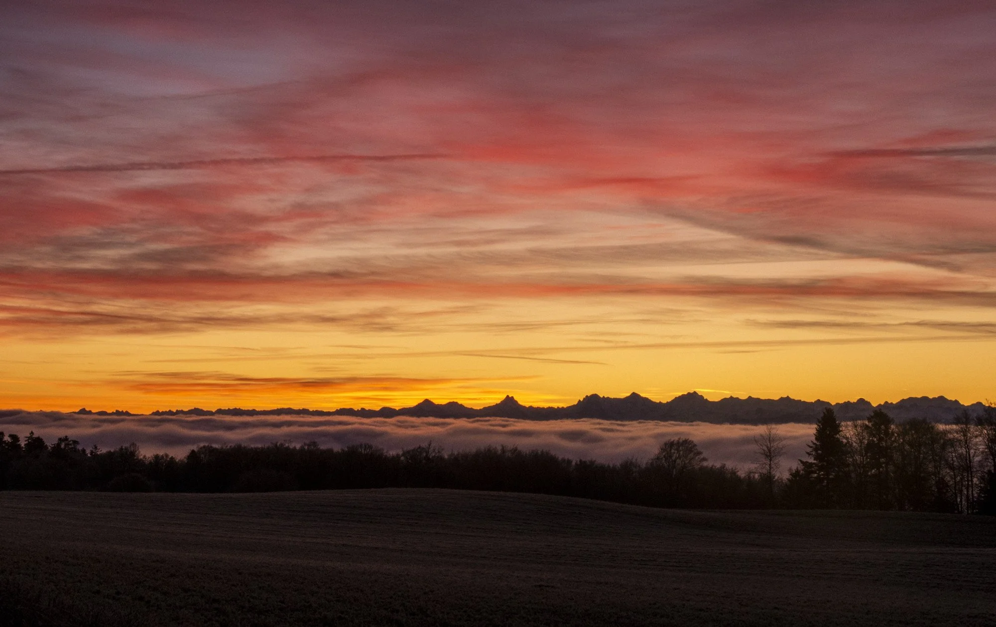 Sunset over a mountain range with colorful orange, pink, and purple sky, clouds, and silhouetted trees and fields in the foreground.
