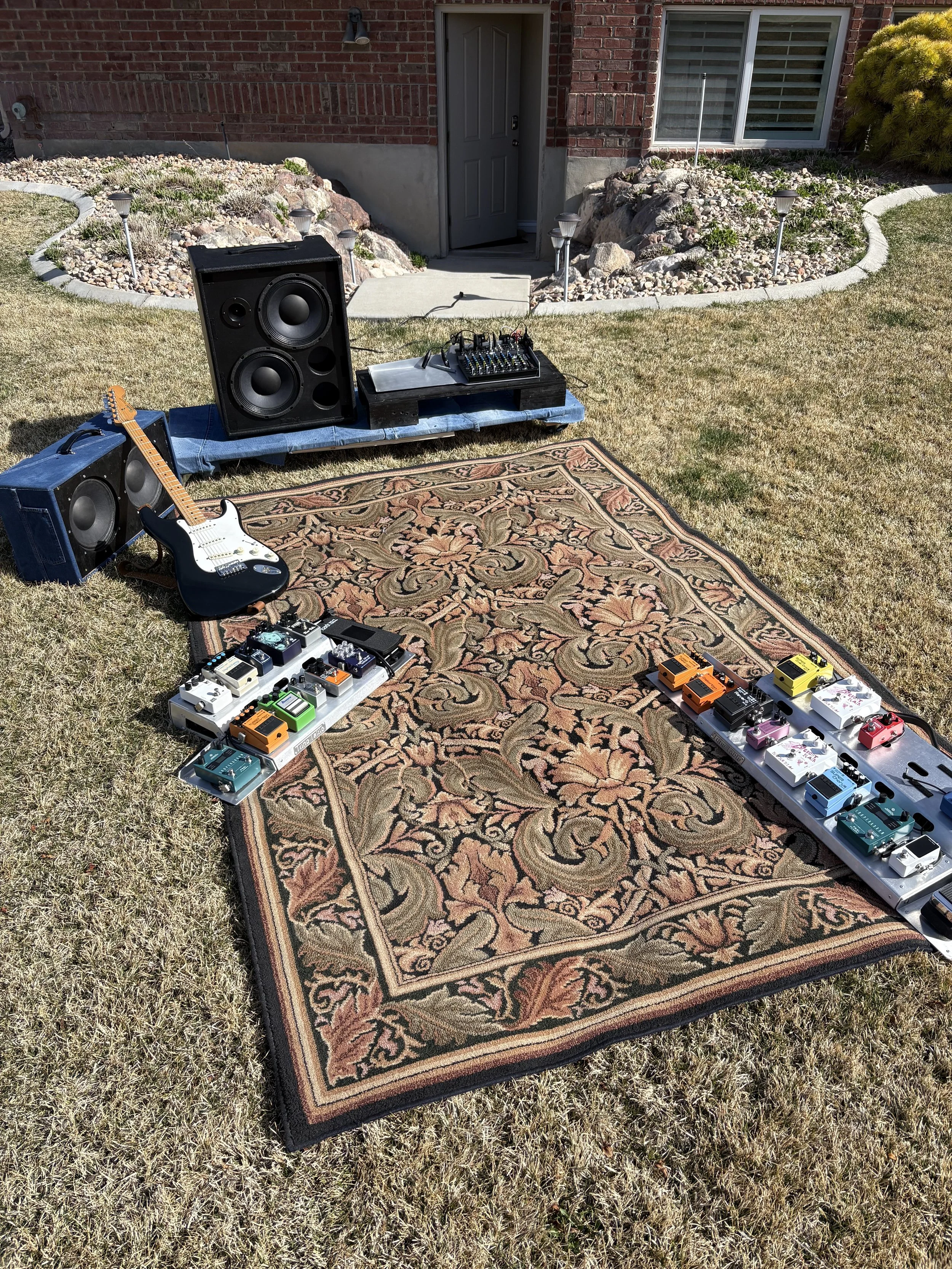Guitar, sound equipment, and effects pedals set up outdoors on a lawn, with a large speaker, smaller speaker, audio mixer, and a patterned rug, positioned near a house with a brick wall, door, and window.