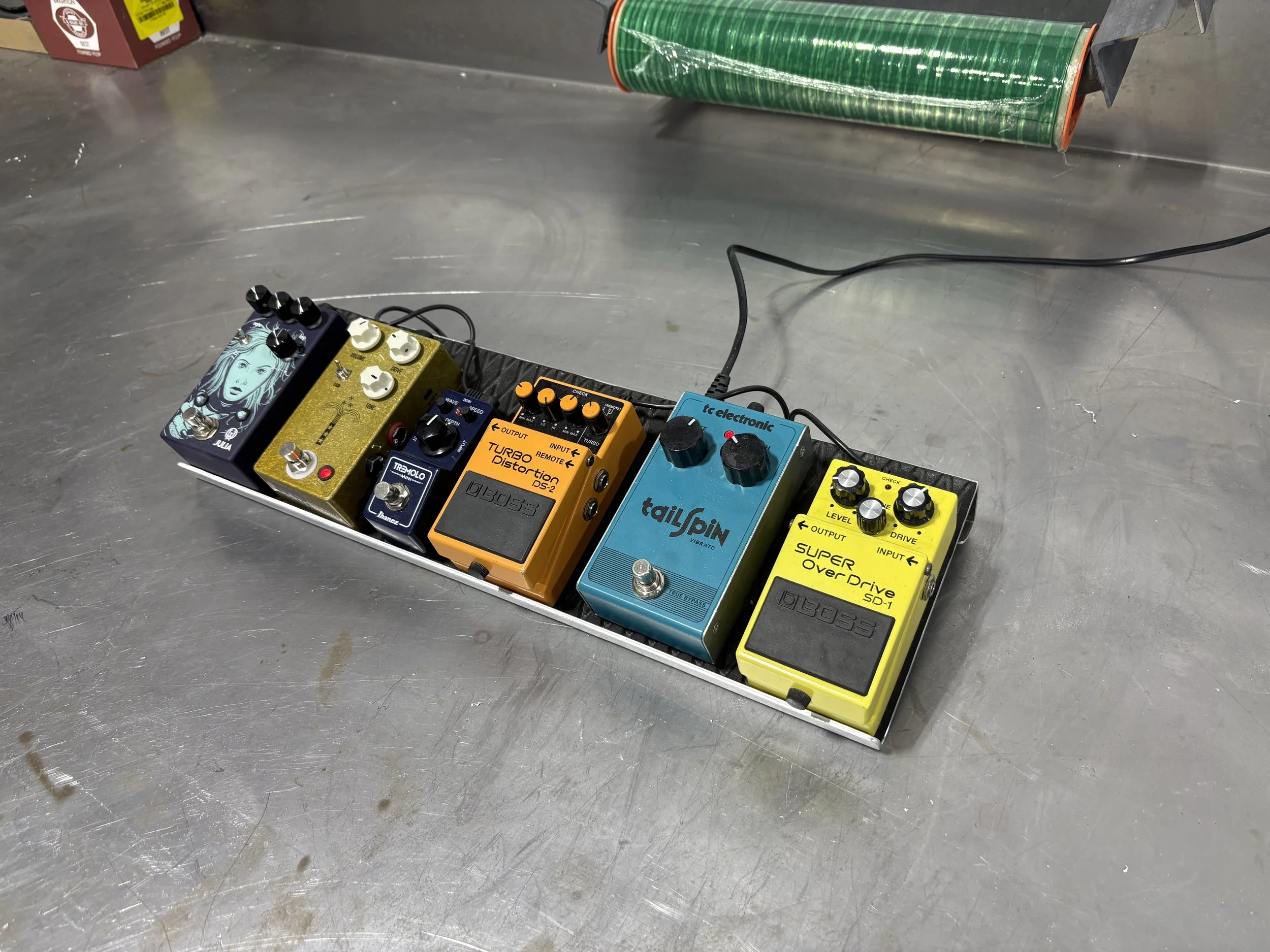 A series of five guitar effect pedals connected on a metal surface, including a custom art pedal, a white and gold pedal, a blue Boss pedal, an orange Boss distortion pedal, and a yellow Boss overdrive pedal. There is also a green roll of tape mounte