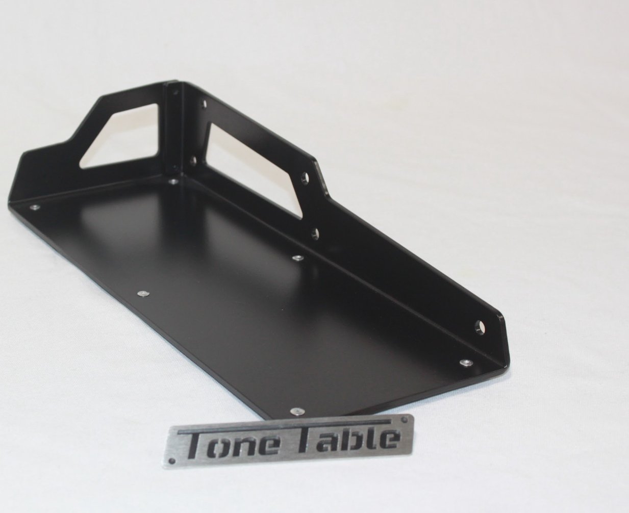 Black metal shelf with four screw holes, placed on a white surface with a metal label reading 'Tone Table'.
