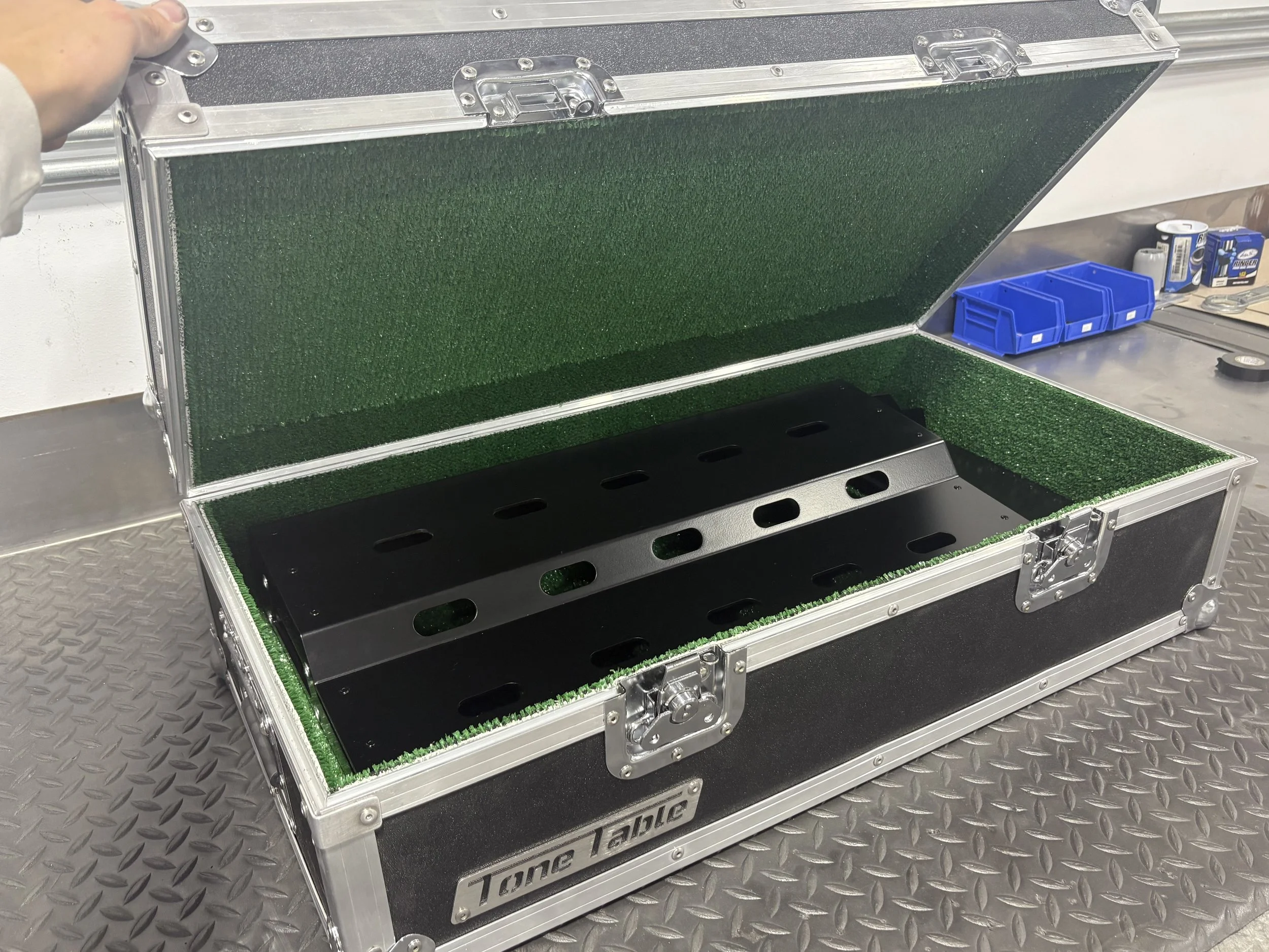 Open black equipment case labeled 'Tone Tackle' with green foam lining, containing metallic black mounting brackets, on a metallic workbench with blue storage bins and tools in the background.