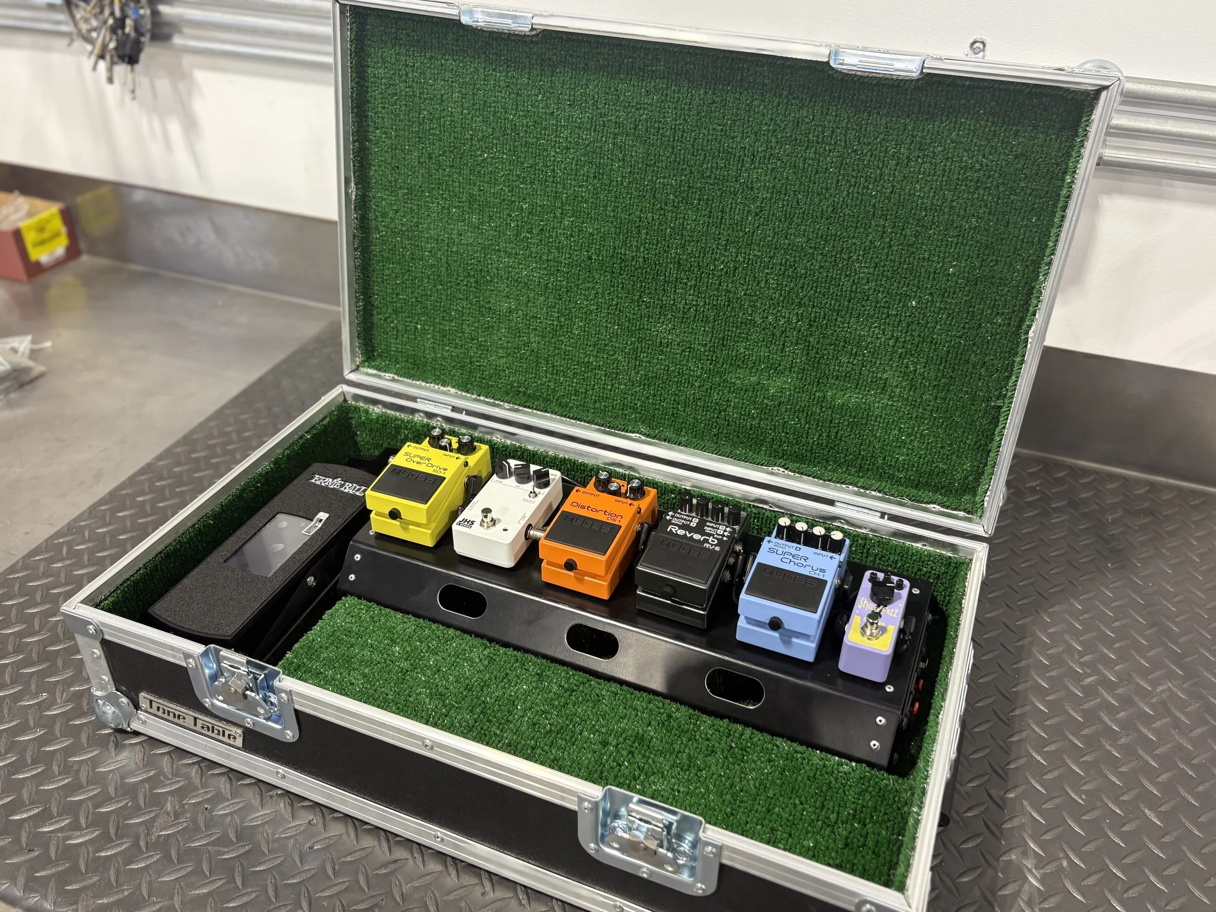 A silver guitar pedal case containing six colorful guitar effect pedals on a black metal pedalboard with green foam padding inside the case.