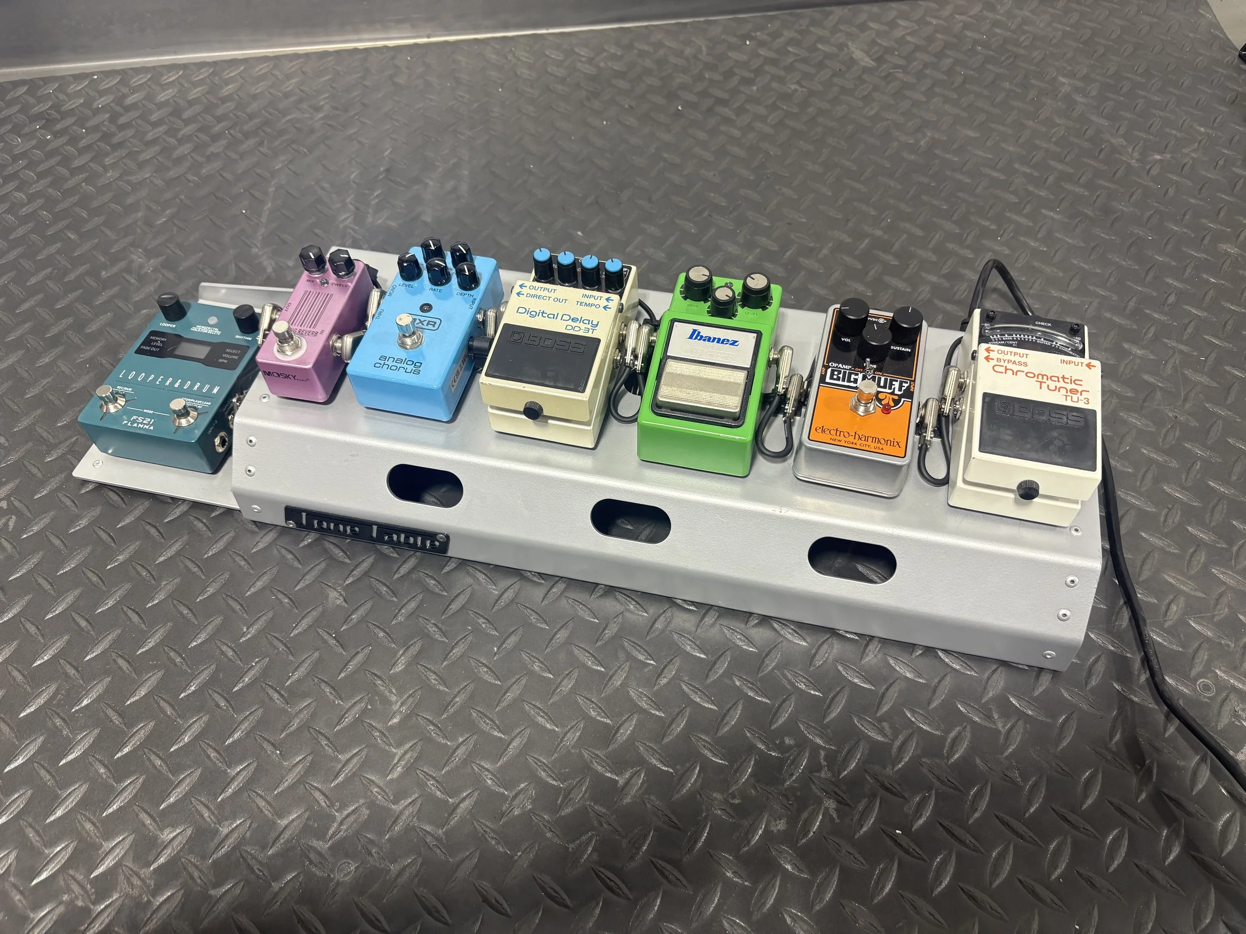 A guitar pedalboard with seven different effects pedals on a metal stand, placed on a textured metal surface.