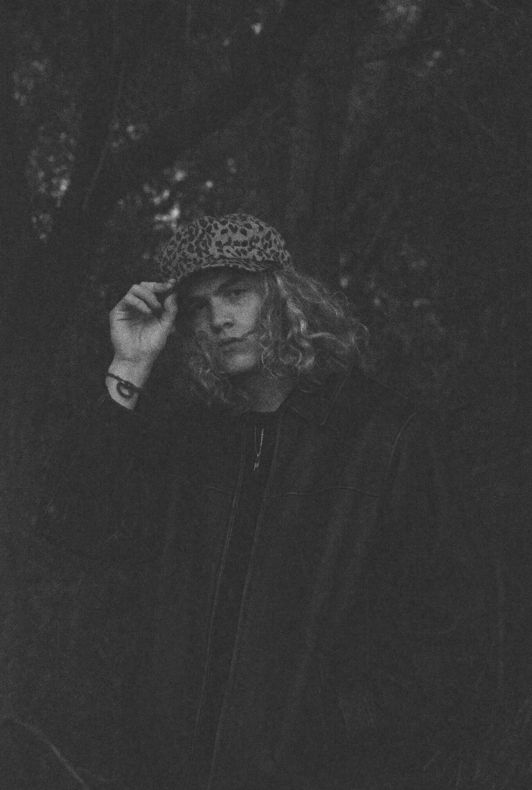 Black and white photo of a woman with curly hair wearing a leopard print hat and a dark jacket, posing outdoors at night.