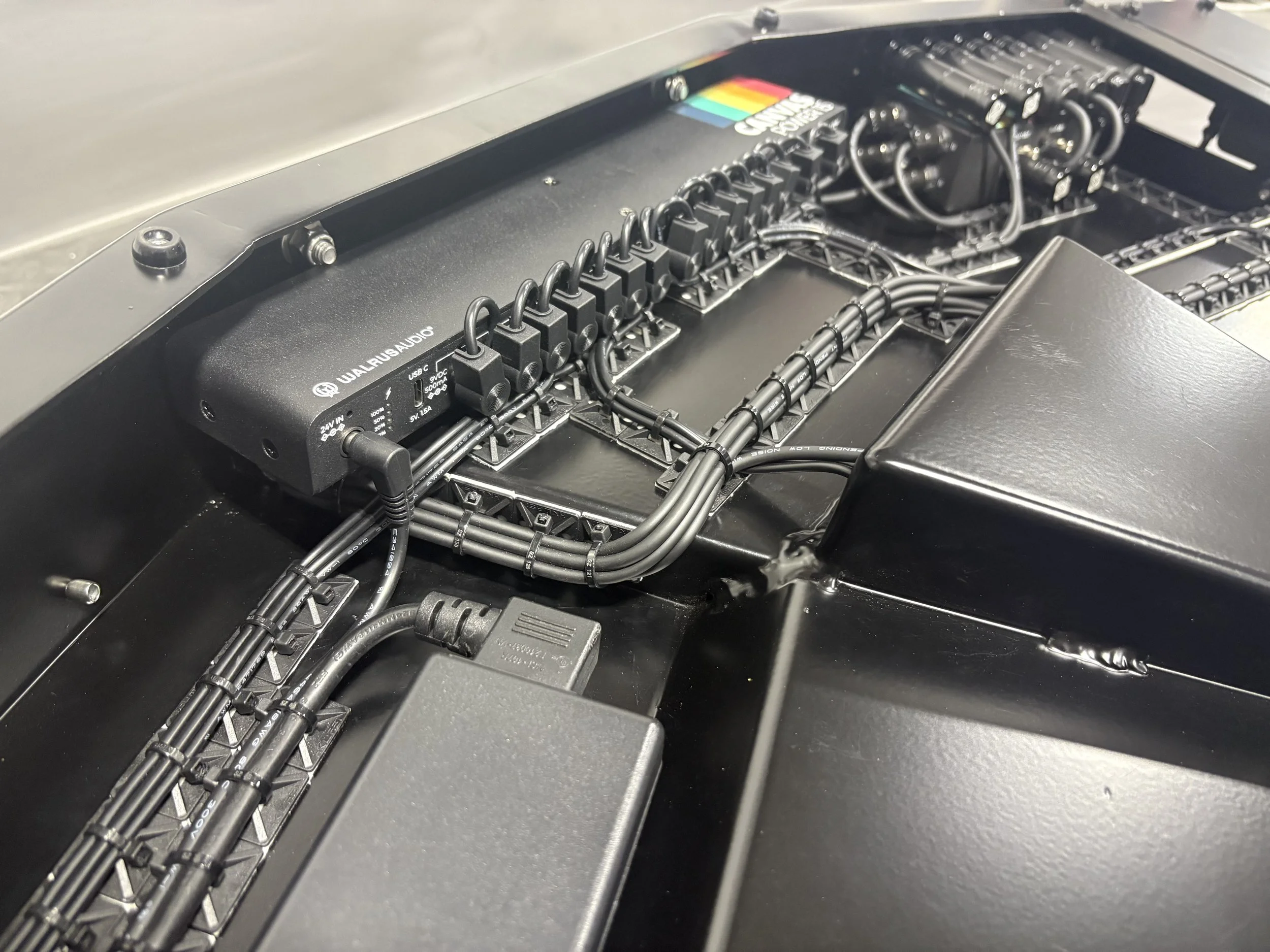 Interior view of a racing car's electronics and wiring, showing a black control unit labeled 'WALRUSH AUDIO' and various cables organized within the black chassis.