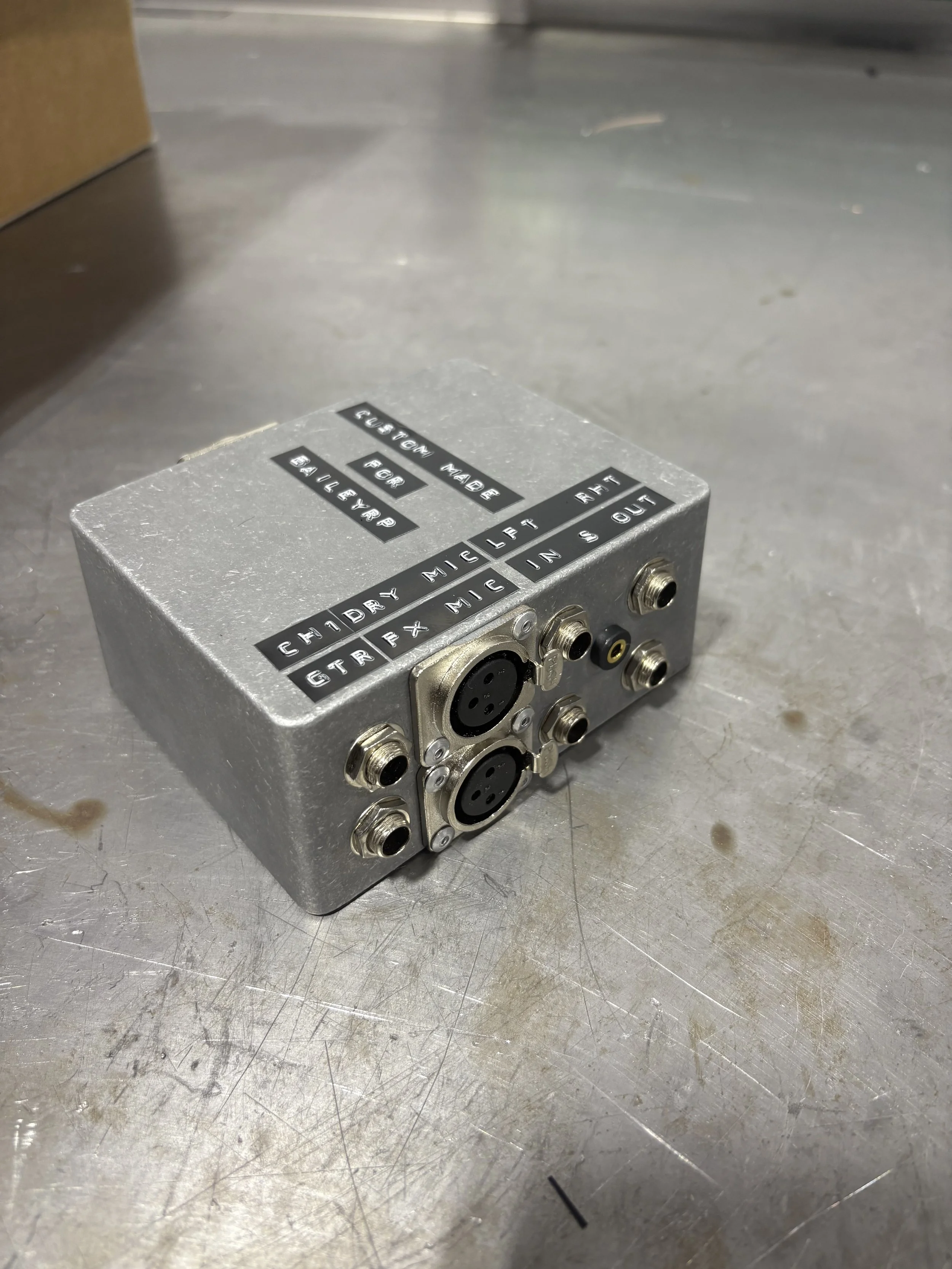 A metallic device with multiple circular input/output ports and black labels. The labels include instructions such as 'CUSTOM MADE', 'FOR', 'GUILTY', 'FOR', 'ALEXANDRIE MIC LEFT', 'GTRX NIC IN S OUT', and 'LEFT', 'RIGHT'.